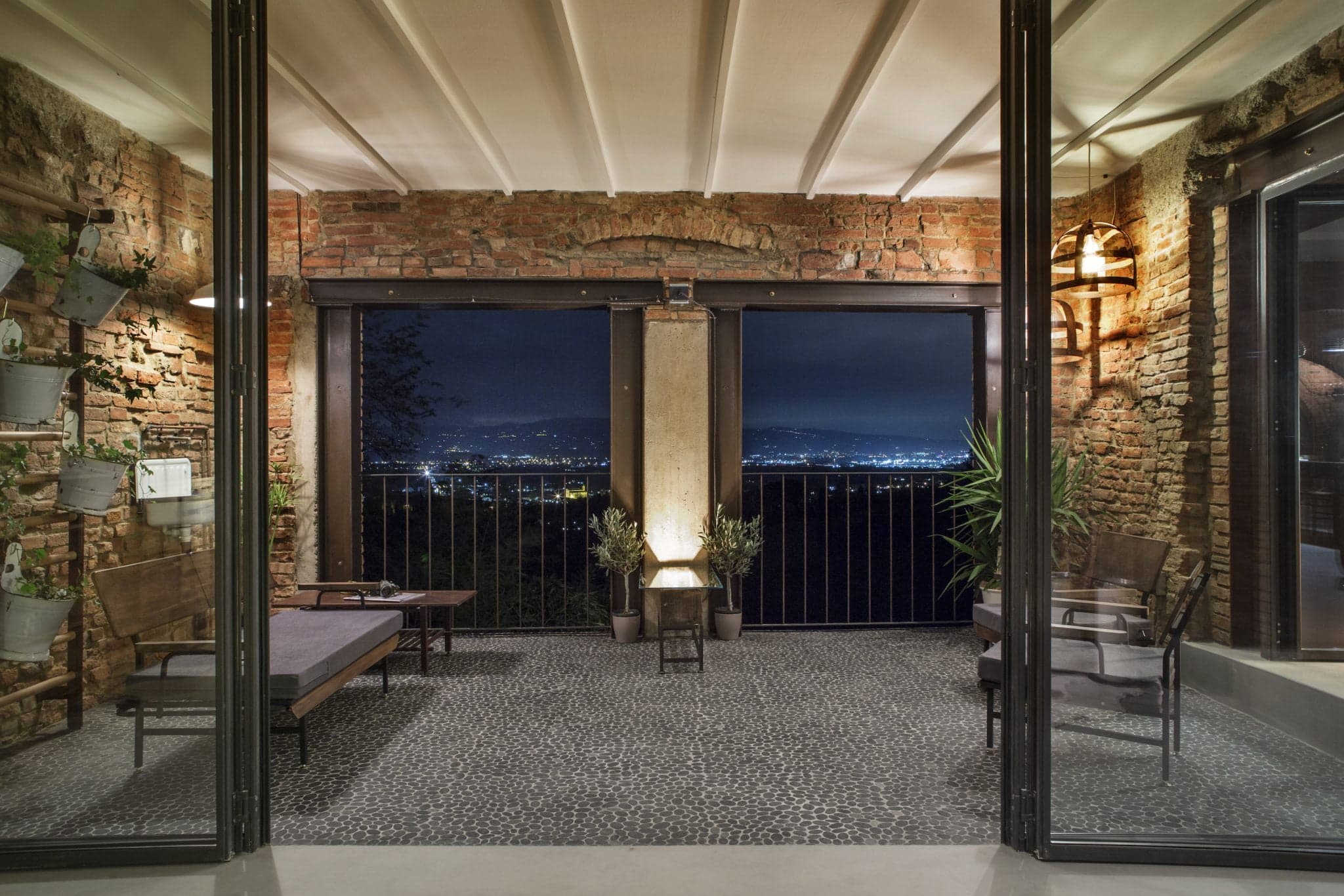 Un balcon rustique et chaleureux avec des murs en briques apparentes, un sol carrelé et des portes vitrées donnant sur un paysage urbain nocturne. L'espace est meublé de chaises en bois, de plantes en pot et d'une petite table, éclairée par des luminaires chaleureux.