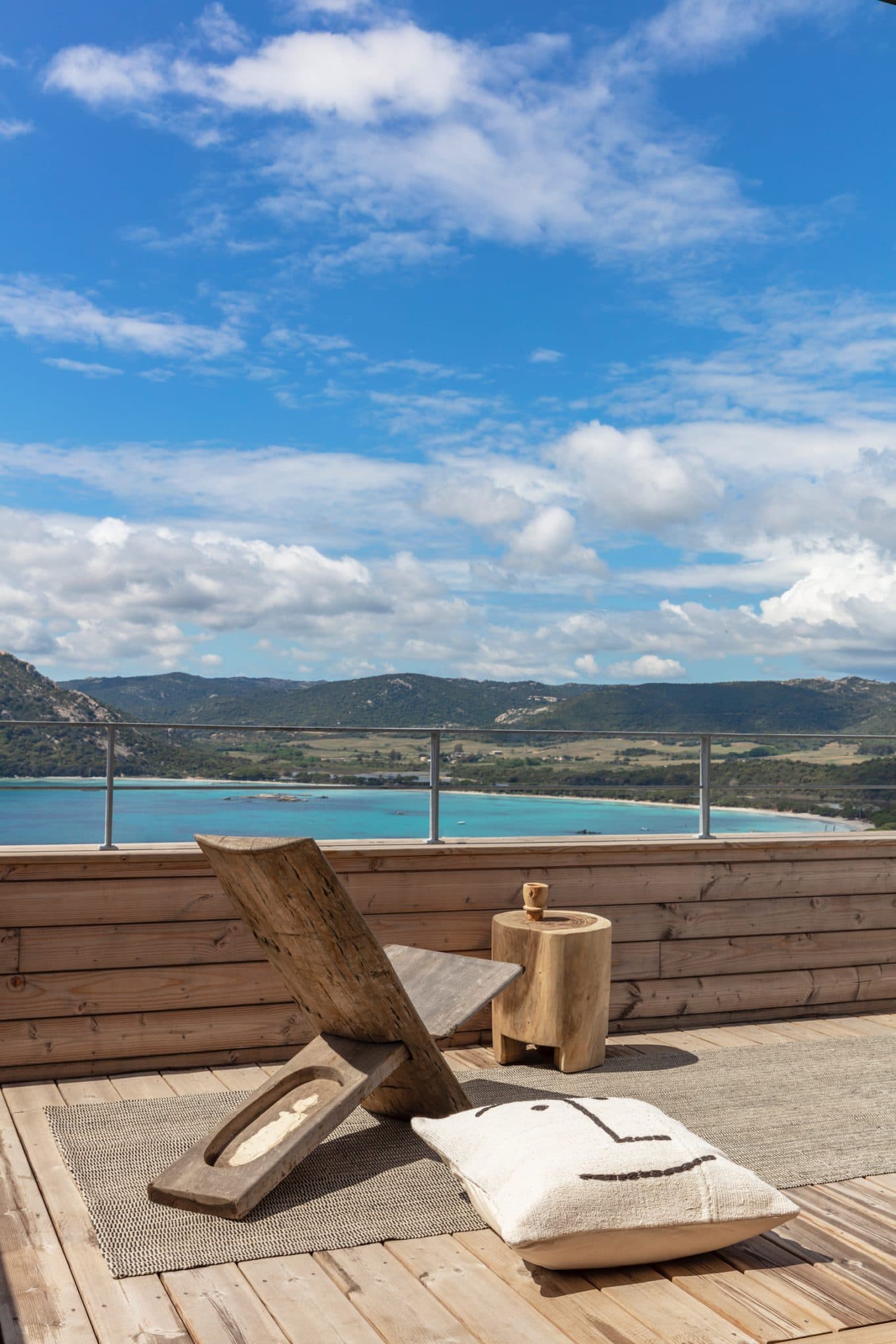 A rooftop wooden terrace featuring an elegant rustic wooden lounge chair and a cylindrical side table. A white cushion with a simple black design rests on a woven rug. The background showcases a breathtaking view of a blue ocean and lush green hills under a partly cloudy sky.