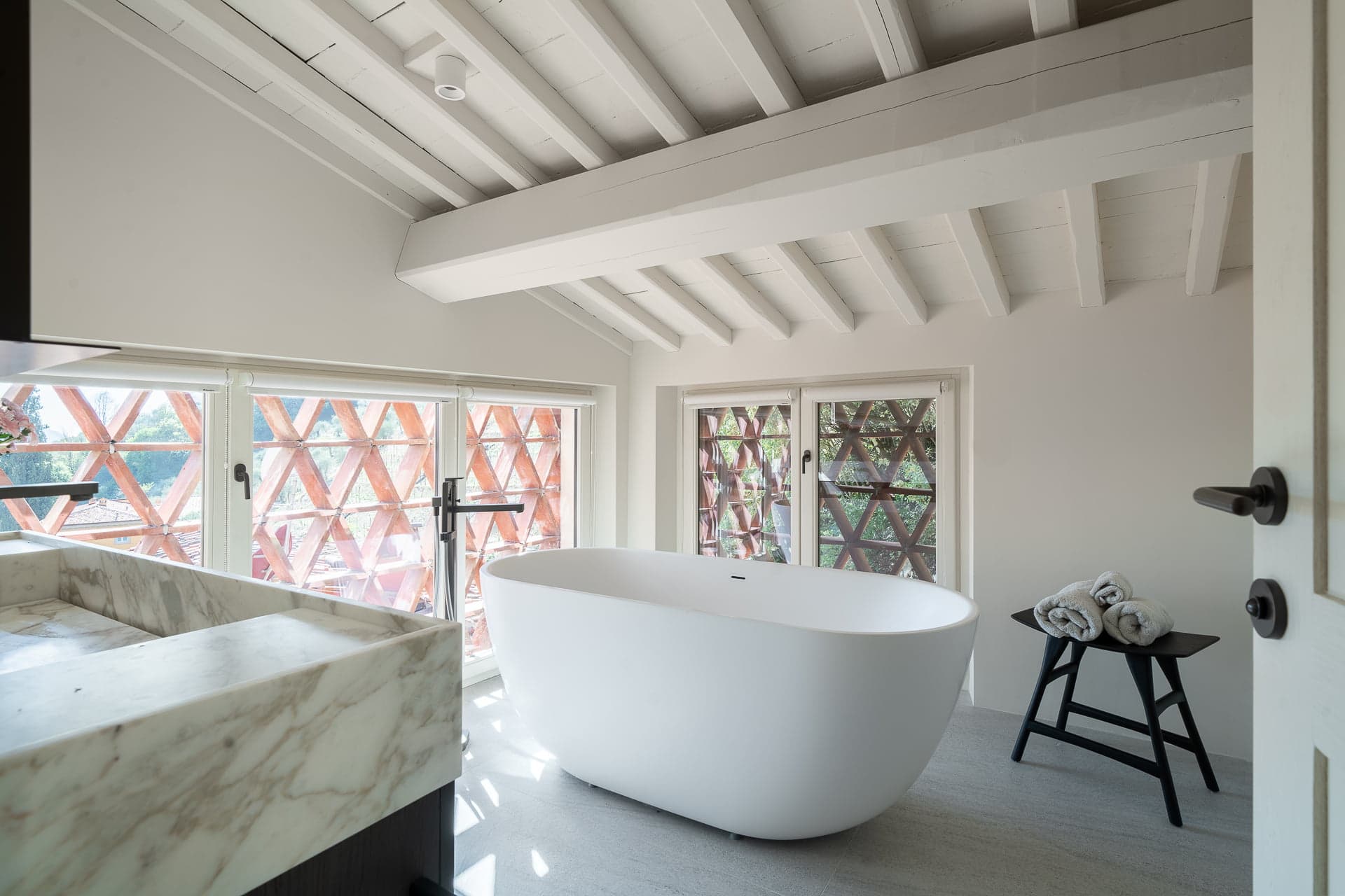A modern bathroom with a white freestanding bathtub positioned near large windows with triangular patterns. The room features a marble countertop, folded towels on a small black stool, and exposed white wooden beams on the ceiling.