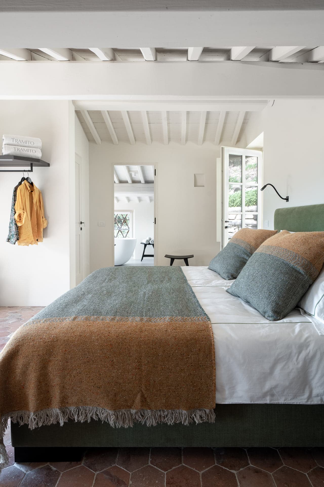 A cozy bedroom features a large bed with green and orange bedding, two pillows, and a blanket. The room has beige walls, a white wooden ceiling, and wooden furniture. A window and a door let in natural light. In the background, a bathroom with white walls and a window is visible.