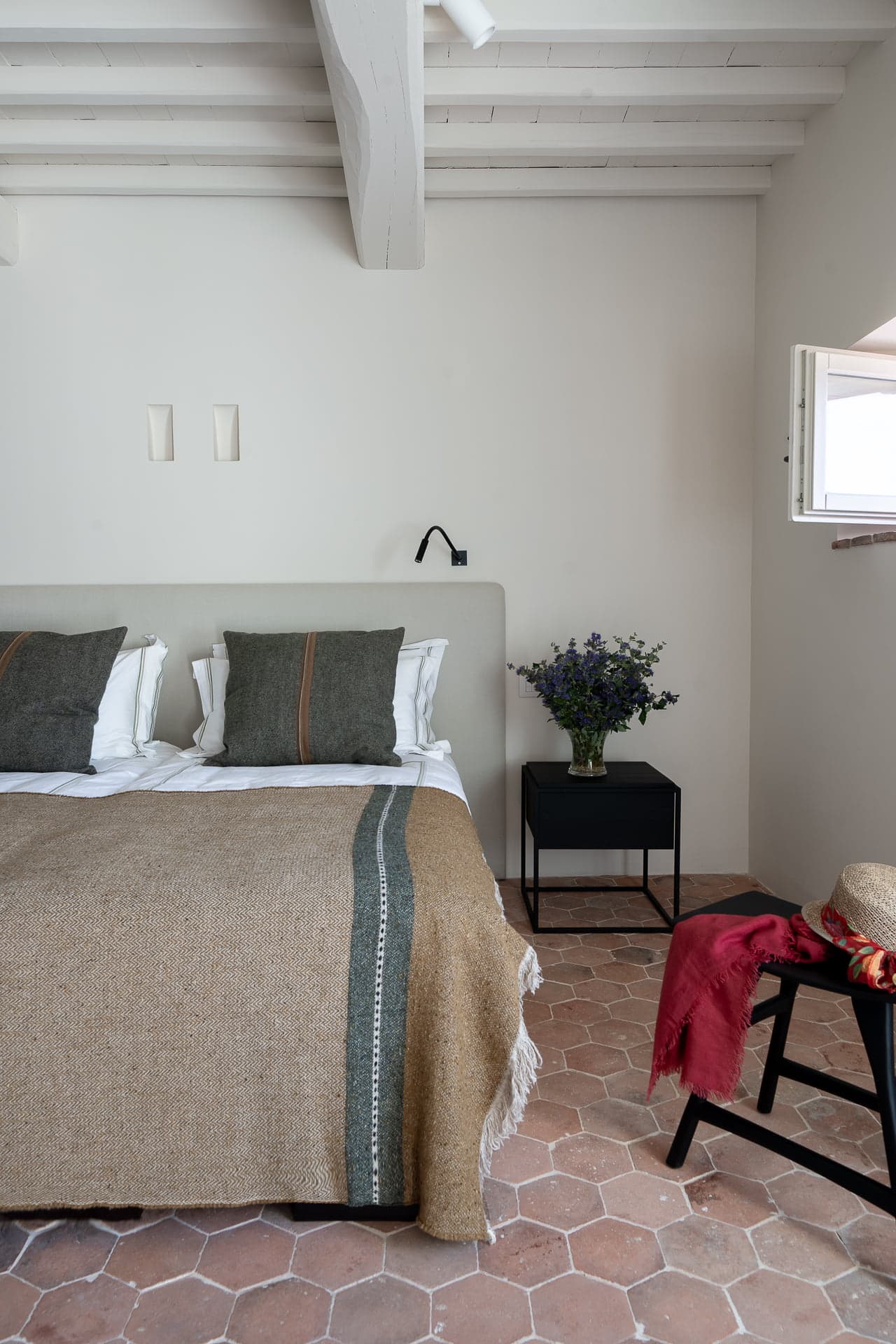 A cozy bedroom features a double bed with a beige headboard and two green pillows. The bed is covered with a beige blanket with green stripes. A black bedside table holds a vase with purple flowers. The room has terracotta tiled flooring and an open window with a white frame.