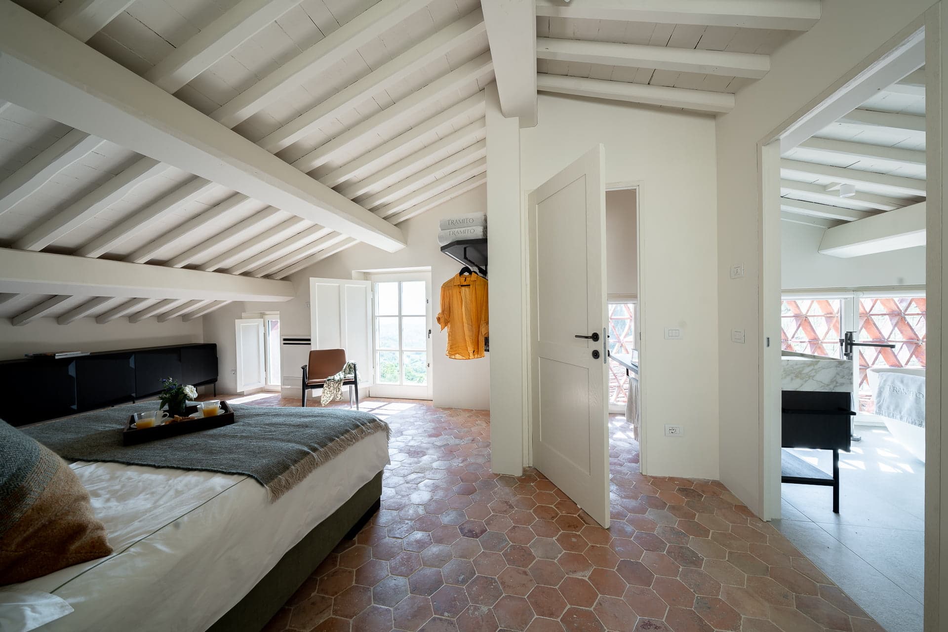 A spacious bedroom with a high ceiling featuring exposed beams. The room includes a comfortable bed, a cozy chair, and hexagonal tiled flooring. Natural light flows through glass doors leading to a balcony. A hallway connects to another room with large windows.