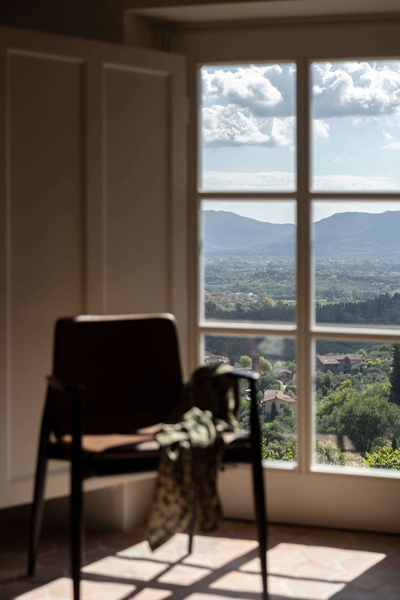 A wooden chair with a piece of green fabric draped over its back sits near a large window. Through the window, there is a panoramic view of a vast landscape with rolling hills, greenery, and clouds under a blue sky. Light spills into the room, casting shadows.