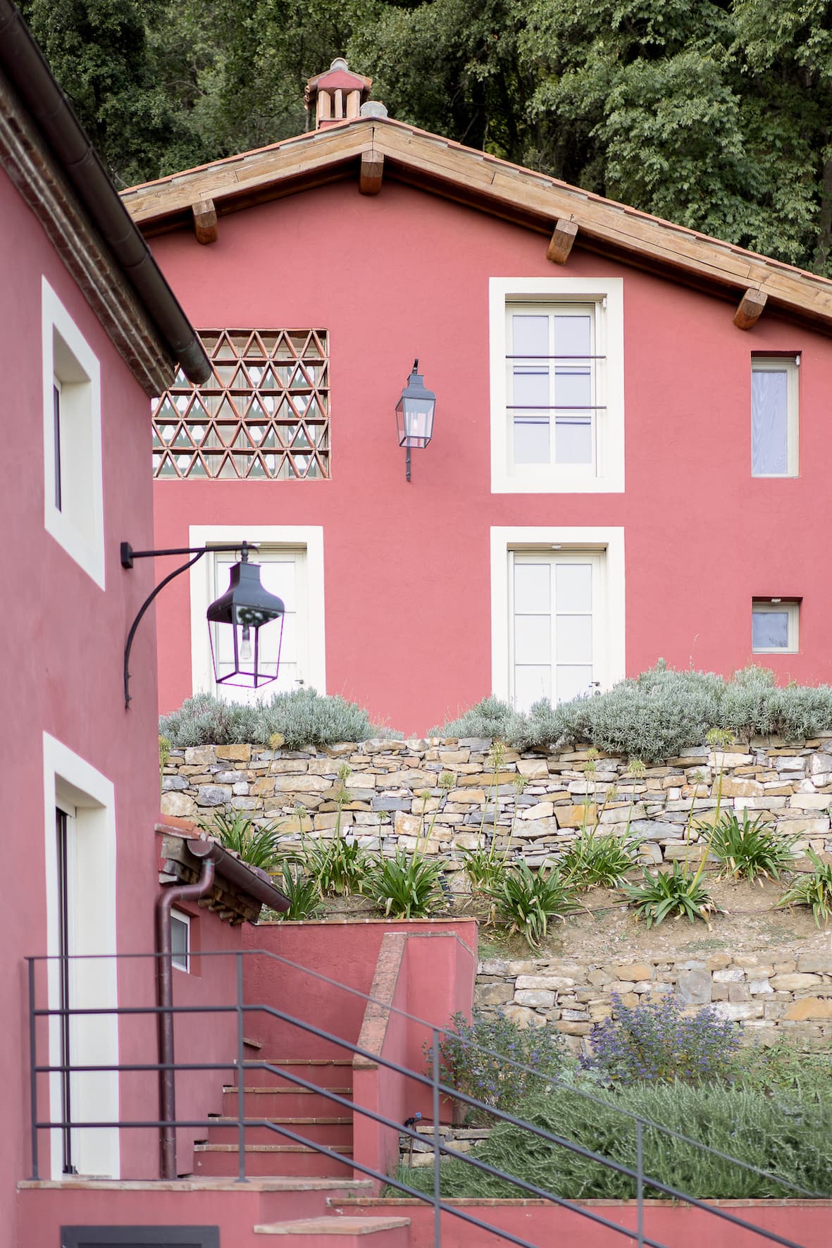 A bright red house with a terracotta roof stands on a lush hillside. The house features a quaint lattice window, decorative outdoor lanterns, and steps leading to a stone retaining wall adorned with green plants and lavender bushes. A smaller red building is in the foreground.