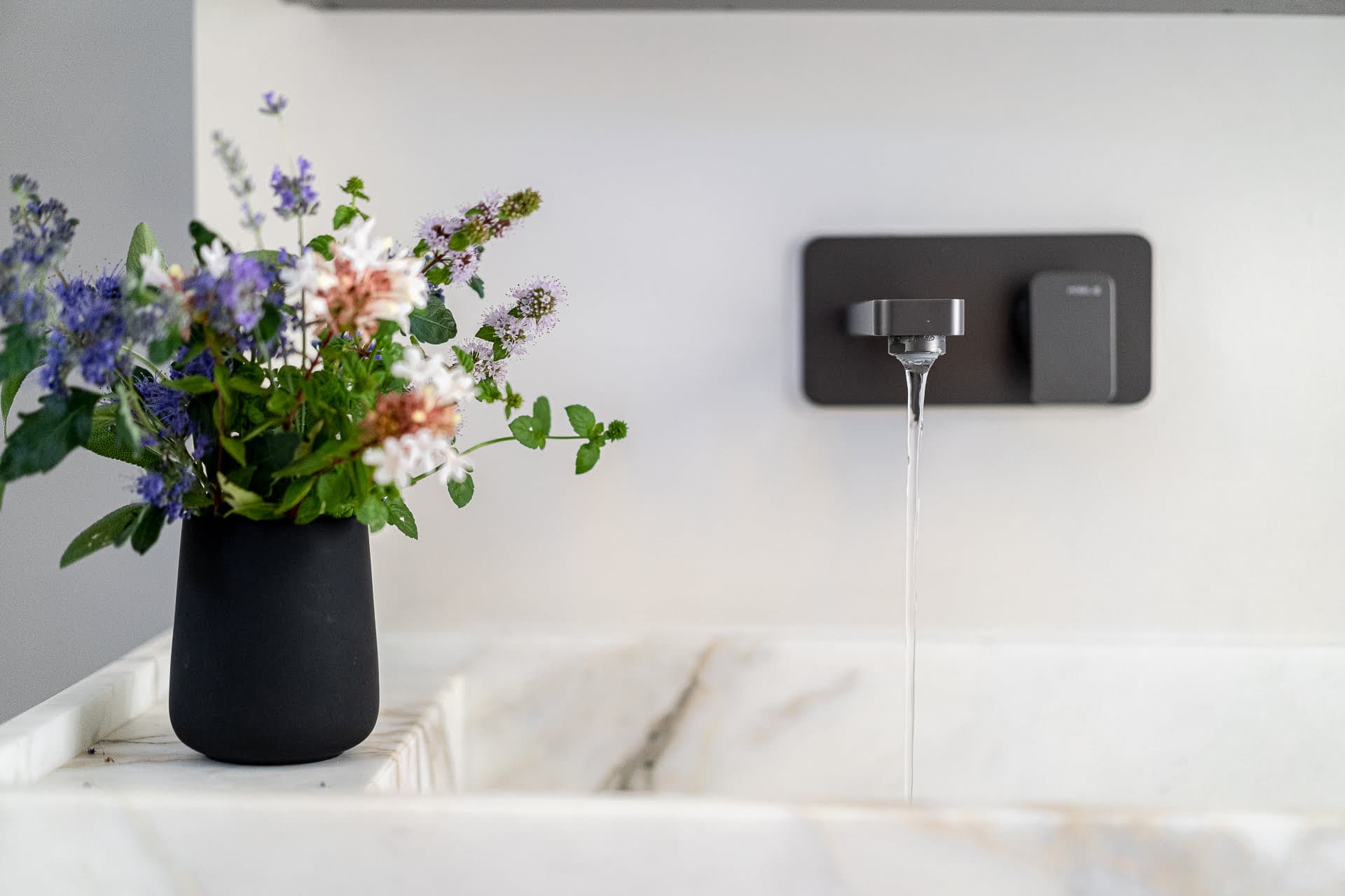 A modern bathroom sink with a sleek wall-mounted faucet is visible, positioned right in the center of a pool of water. To the left of the sink, a black vase holds a colorful arrangement of flowers, adding a touch of natural beauty to the contemporary space.