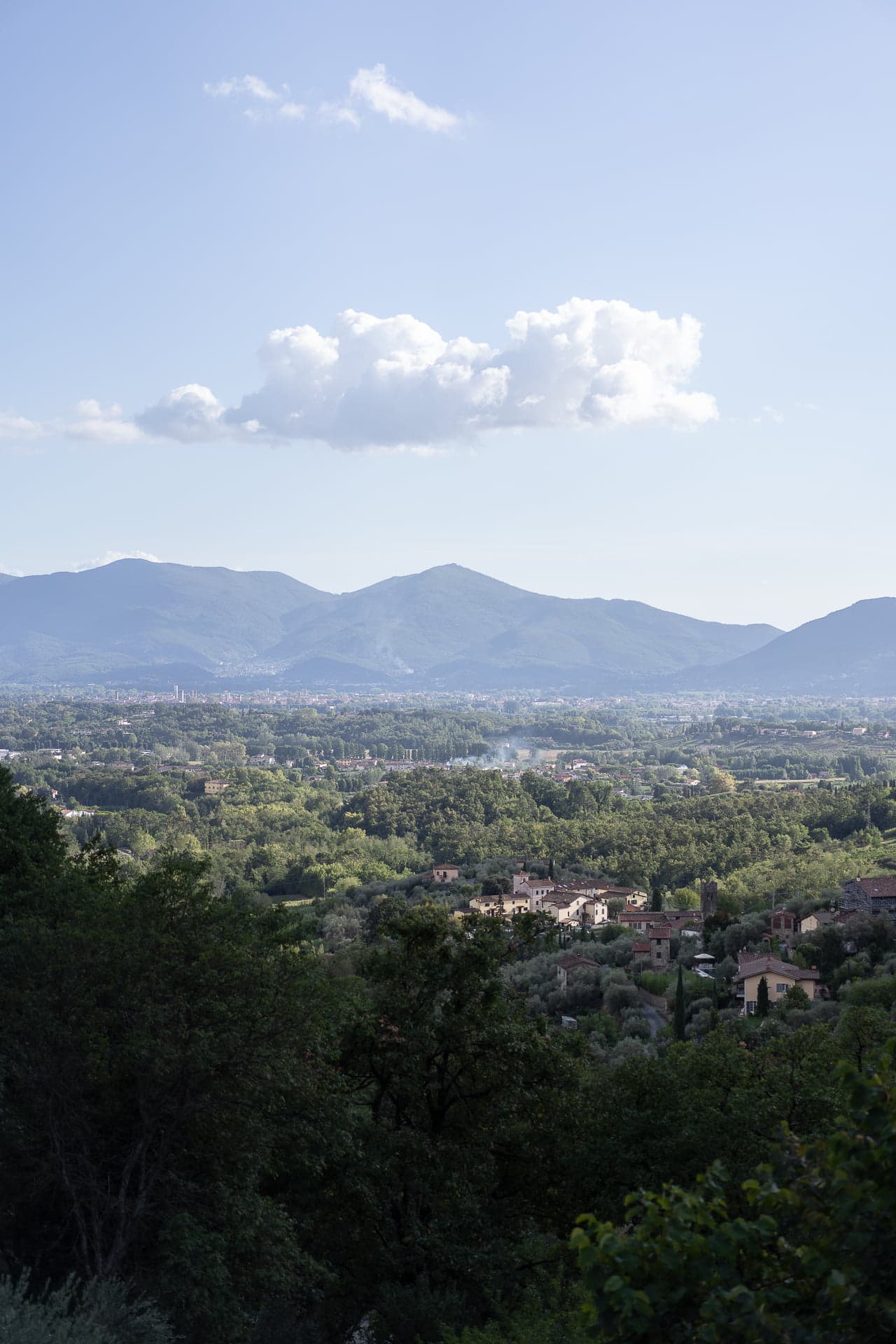 A serene landscape depicts a valley with clusters of houses nestled among lush green trees. In the background, misty mountains rise under a clear blue sky with a few fluffy clouds floating above. The scene evokes a sense of peace and tranquility.
