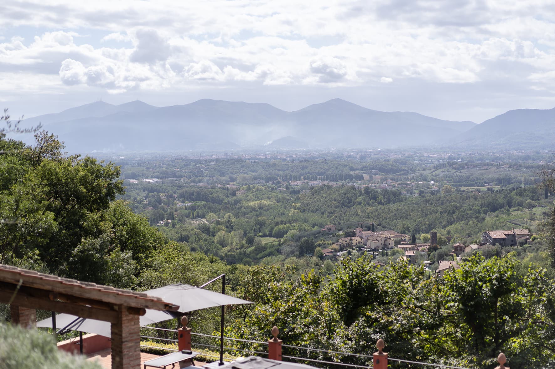 A panoramic view of a verdant and lush valley, dotted with houses and buildings, framed by distant mountains under a partly cloudy sky. In the foreground, part of a building's roof and an outdoor umbrella are visible.