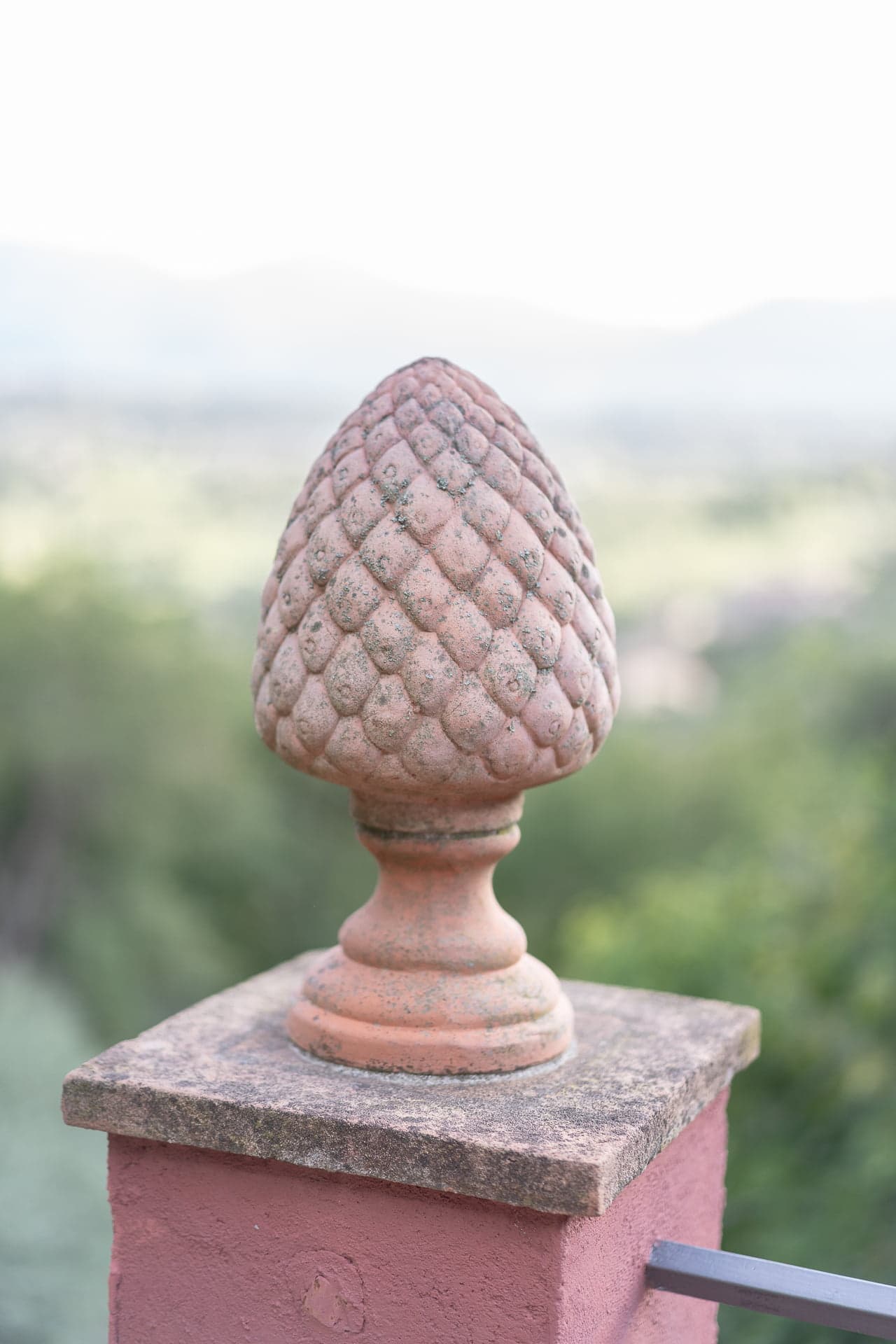 A terracotta sculpture depicting a pinecone rests on a pedestal atop a brick column, with a blurred natural landscape in the background. The soft focus highlights the detailed texture of the sculpture.