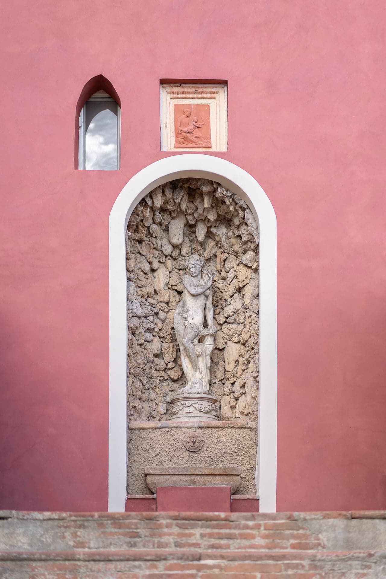 A pink wall features a recessed alcove housing a white stone sculpture of a nude female figure. Above the sculpture, there is a small square relief artwork. The alcove arch and a nearby window are framed in white. Steps lead up to the alcove.