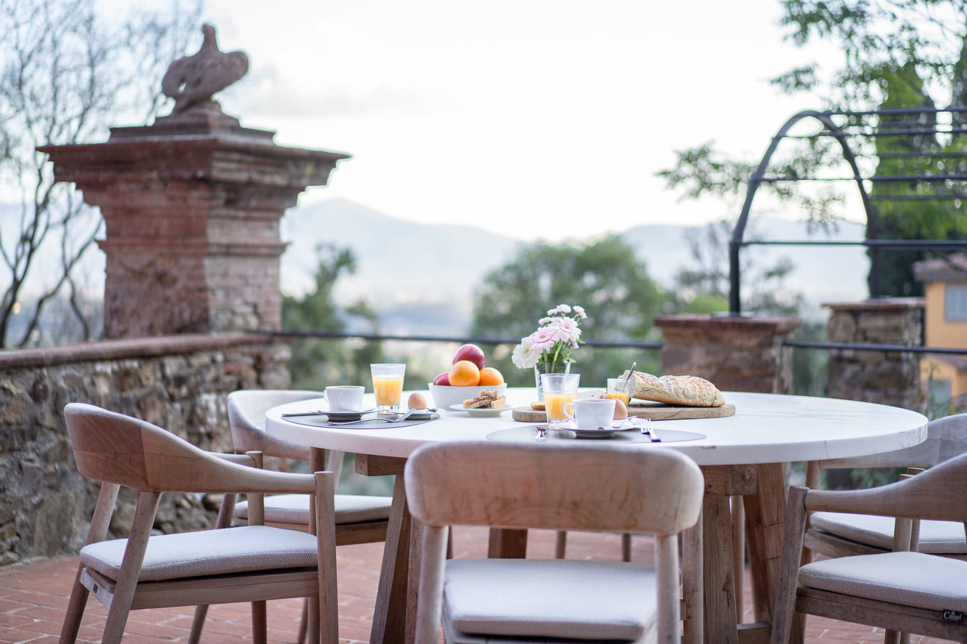 A white round table set for breakfast on a terrace with four wooden chairs. The table is arranged with plates of fruit, glasses of juice, a loaf of bread, coffee cups, and a vase with flowers. The terrace overlooks a panoramic view of trees and mountains.