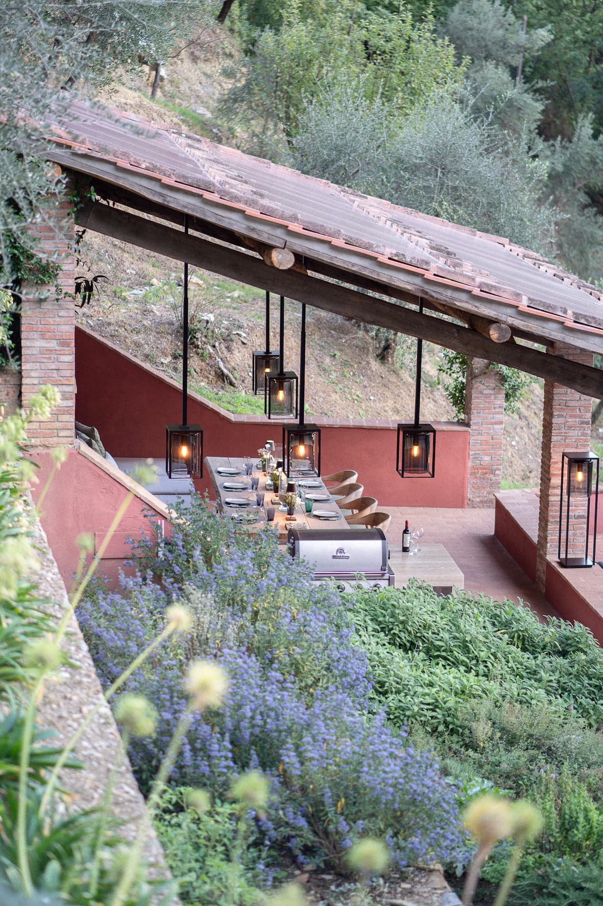 An outdoor dining area is set up under a rustic covered patio with brick columns. The table is elegantly set with dishware and surrounded by chairs. Pendant lights hang from the wooden ceiling, and lush vegetation surrounds the space, enhancing the serene ambiance.