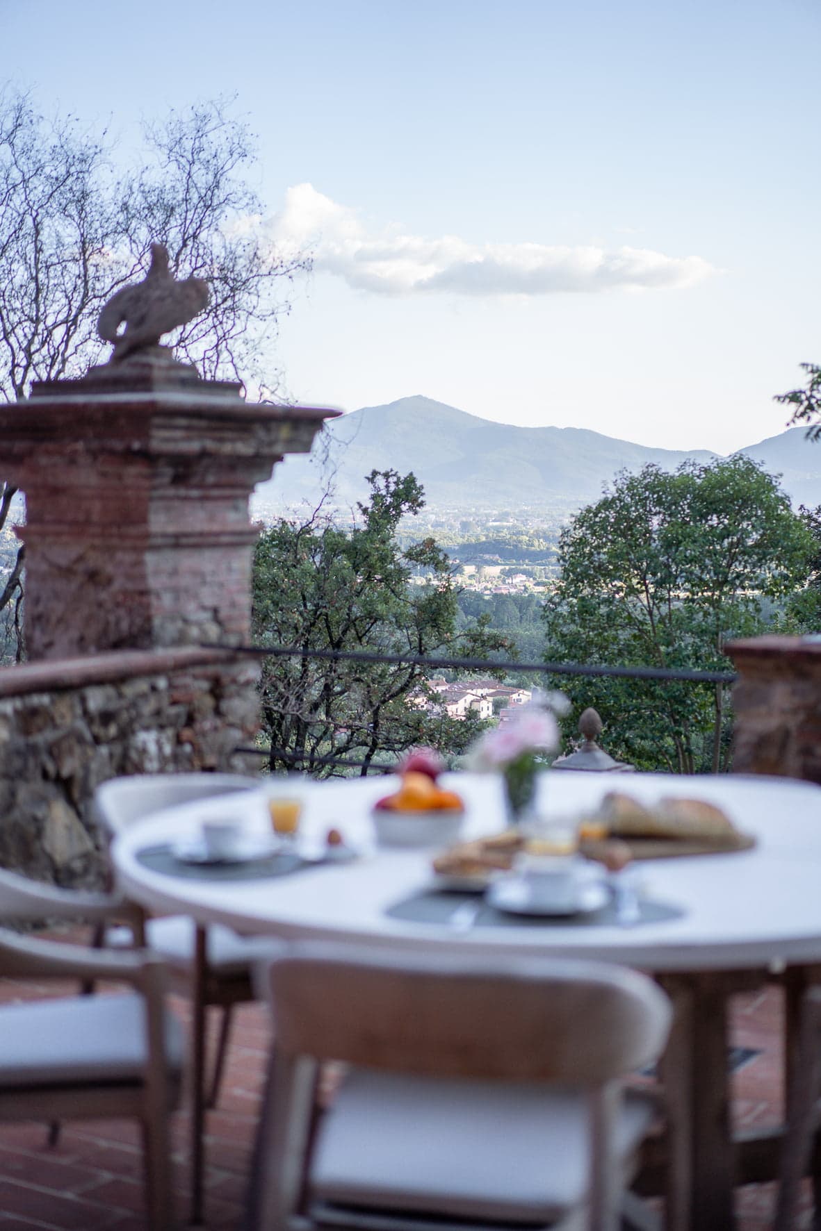 An outdoor dining table set for breakfast, featuring pastries, fruits, and drinks, placed on a terrace with a panoramic view of the mountains in the background. The atmosphere is serene, with lush vegetation and a distant town visible under a clear blue sky.