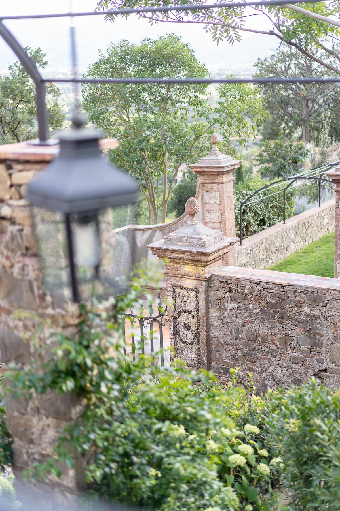 A serene garden scene features a brick and stone wall with ornamental details, a wrought-iron gate, and lush vegetation. A vintage-style lamppost is in the foreground, and trees and distant hills are visible in the background, creating a tranquil atmosphere.