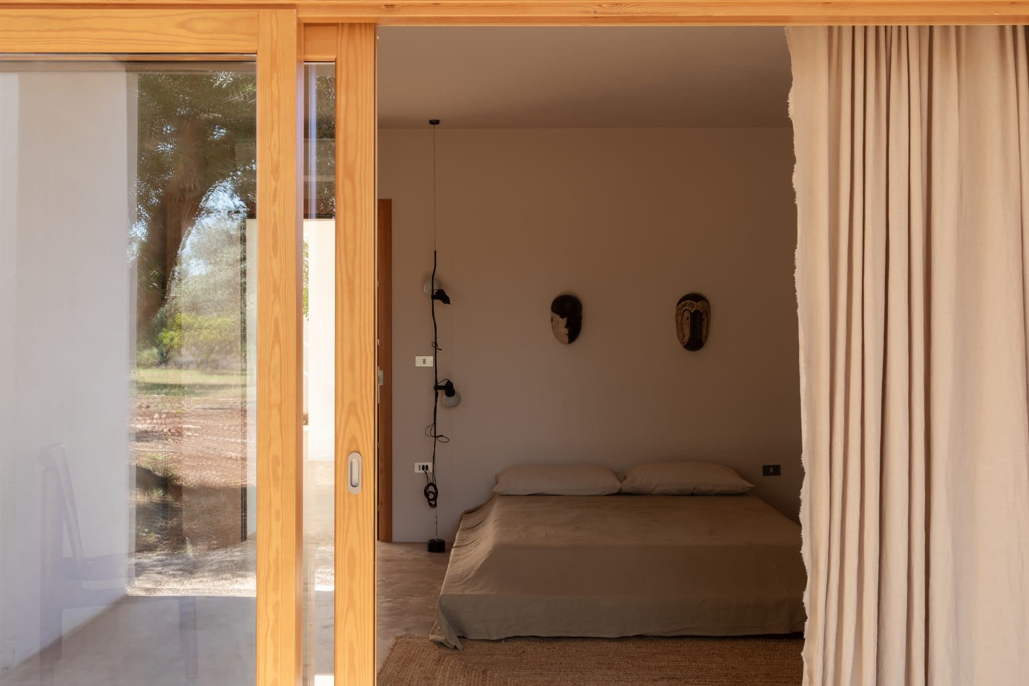 A minimalist bedroom is visible through an open sliding glass door. The room features a low bed with beige sheets, two decorative masks on the wall above, a pendant light hanging from the ceiling, and beige curtains partially drawn to the side. An outdoor tree is visible through the window.