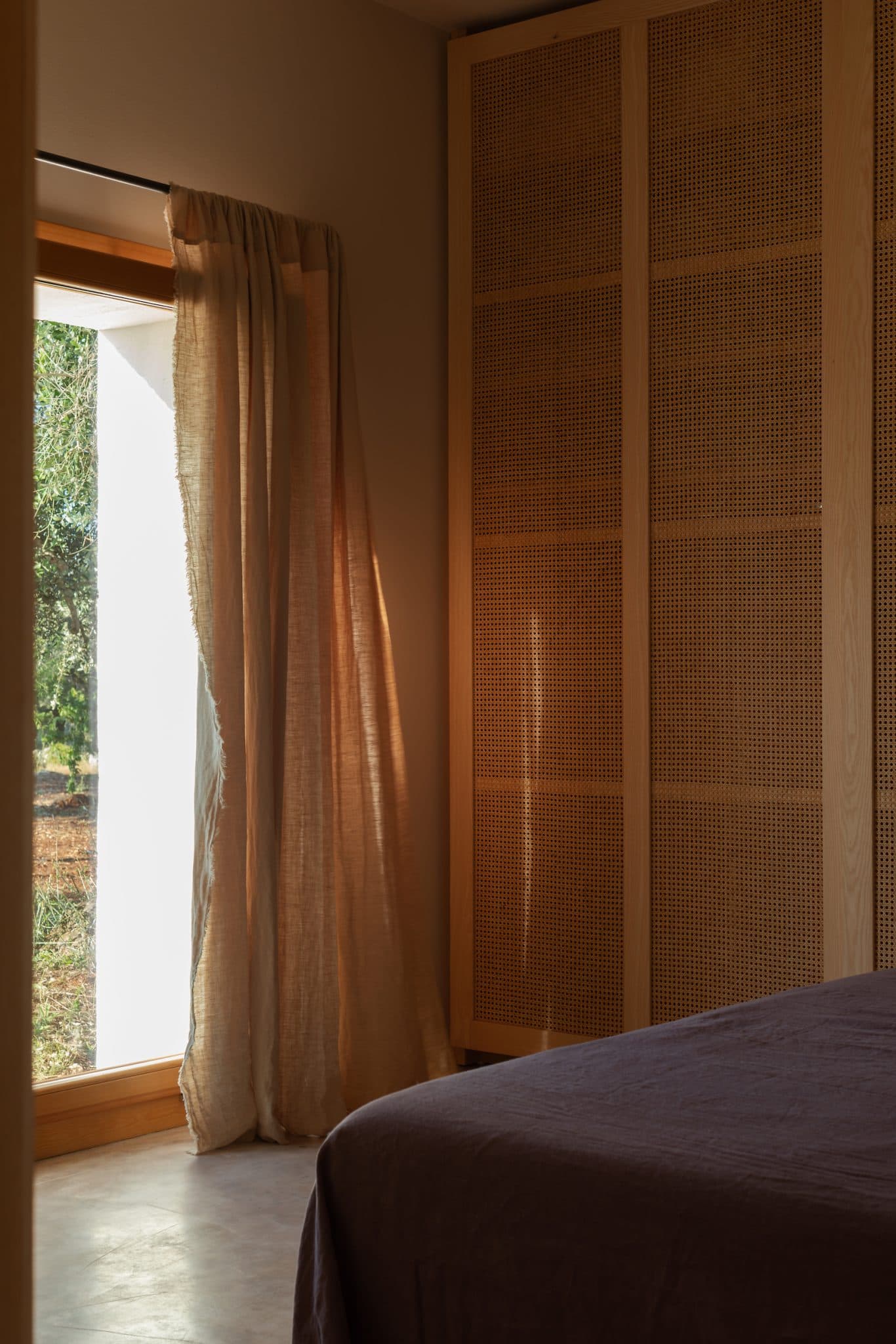 A cozy bedroom features a wooden wardrobe with woven rattan panels and partially translucent curtains that let light filter through a large window. A bed with a dark-colored cover is visible in the foreground, with soft lighting creating a warm atmosphere.