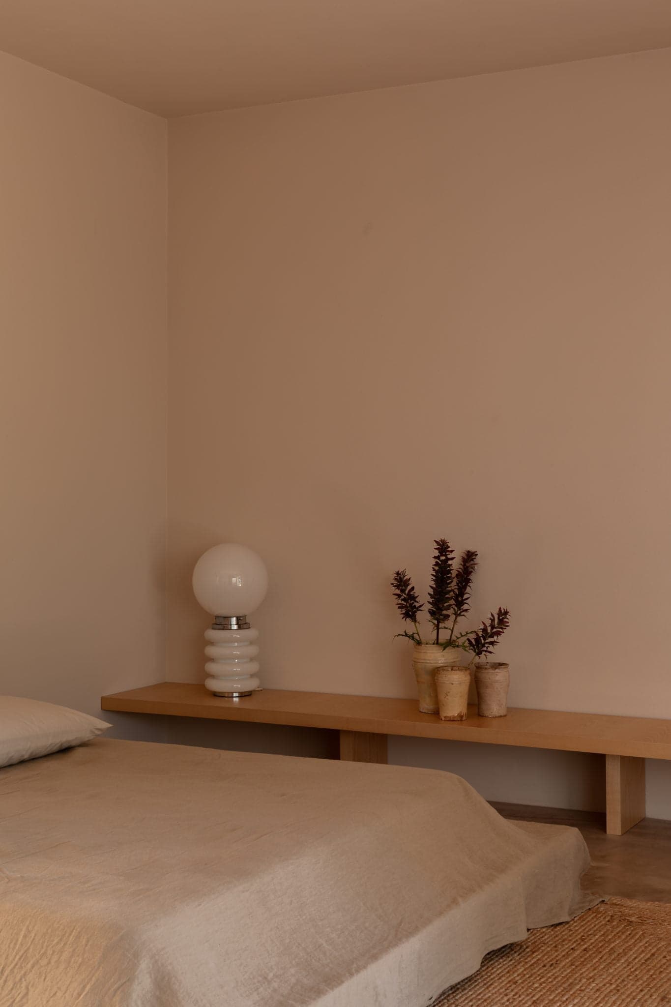 A minimalist bedroom features a beige bed with matching sheets, a wooden bench serving as a nightstand, a round white lamp with a spiral base, and three rustic vases with dried plants. The walls and floor are also in neutral tones, creating a serene atmosphere.