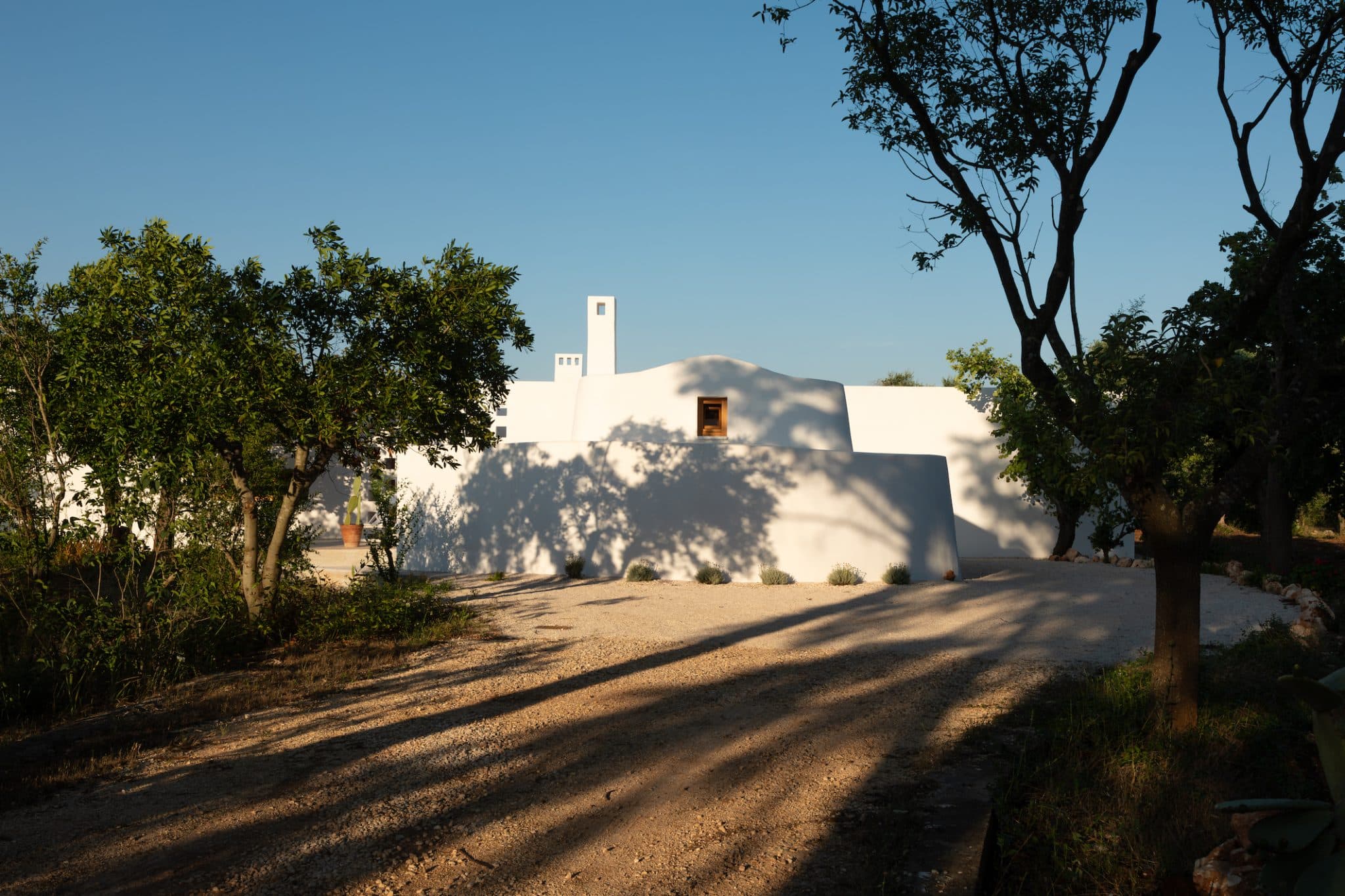 Une maison aux murs blancs au design minimaliste est entourée d'arbres et de verdure. Un chemin de gravier mène à l'entrée, projetant de longues ombres dans la lumière du soleil de fin d'après-midi. Le ciel est clair et bleu, mettant en valeur le cadre paisible et rustique.