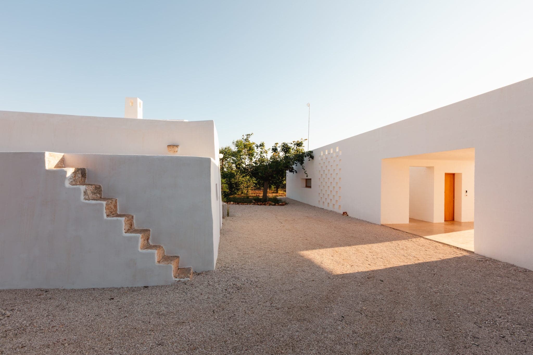 A white, modern, minimalist building with clean lines in an open space. The building features an external staircase leading to an upper area. The surrounding ground is covered with light-colored gravel, and greenery is visible in the background.