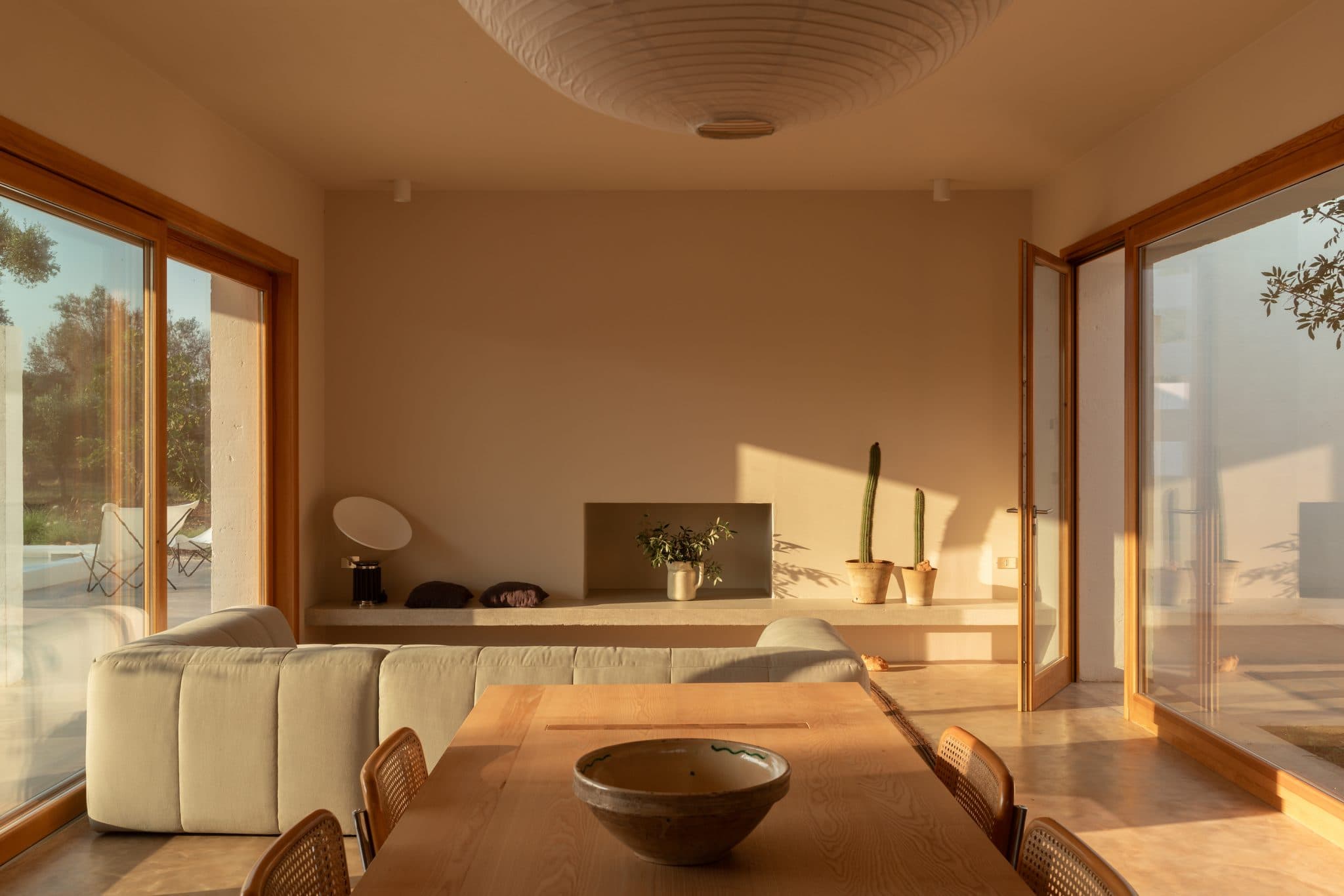 A minimalist living and dining area is bathed in warm natural light, featuring a wooden dining table with a central bowl, a light-colored sofa, and large sliding glass doors. The space is decorated with a table lamp, potted plants, and a neutral color palette.