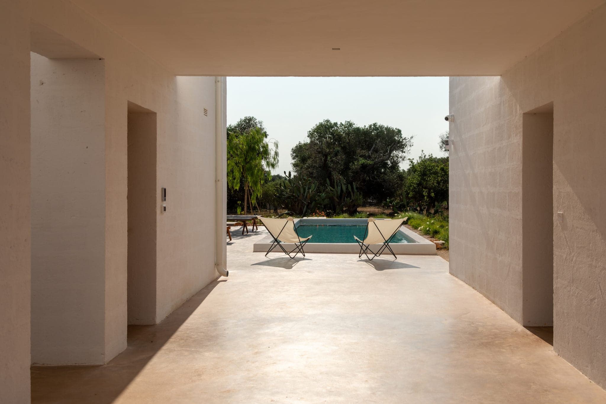 A serene outdoor scene of a minimalist villa shows two lounge chairs positioned in front of a rectangular pool. The pool is surrounded by lush greenery, visible through a large open corridor flanked by white walls under a clear sky.