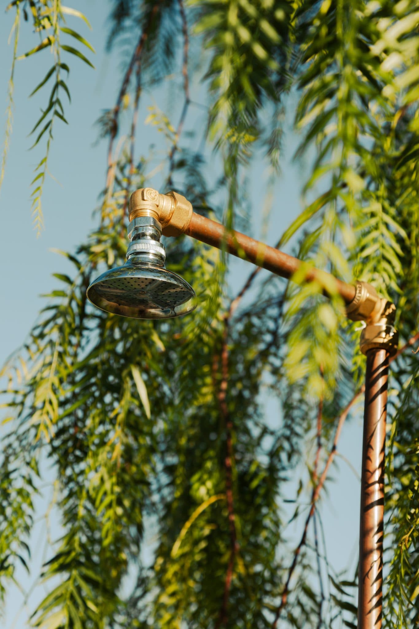 An outdoor shower head is mounted on a copper pipe, with green foliage and trees in the background. The metal shower head features an industrial design, contrasting with the natural landscape. The sky is clear and blue.