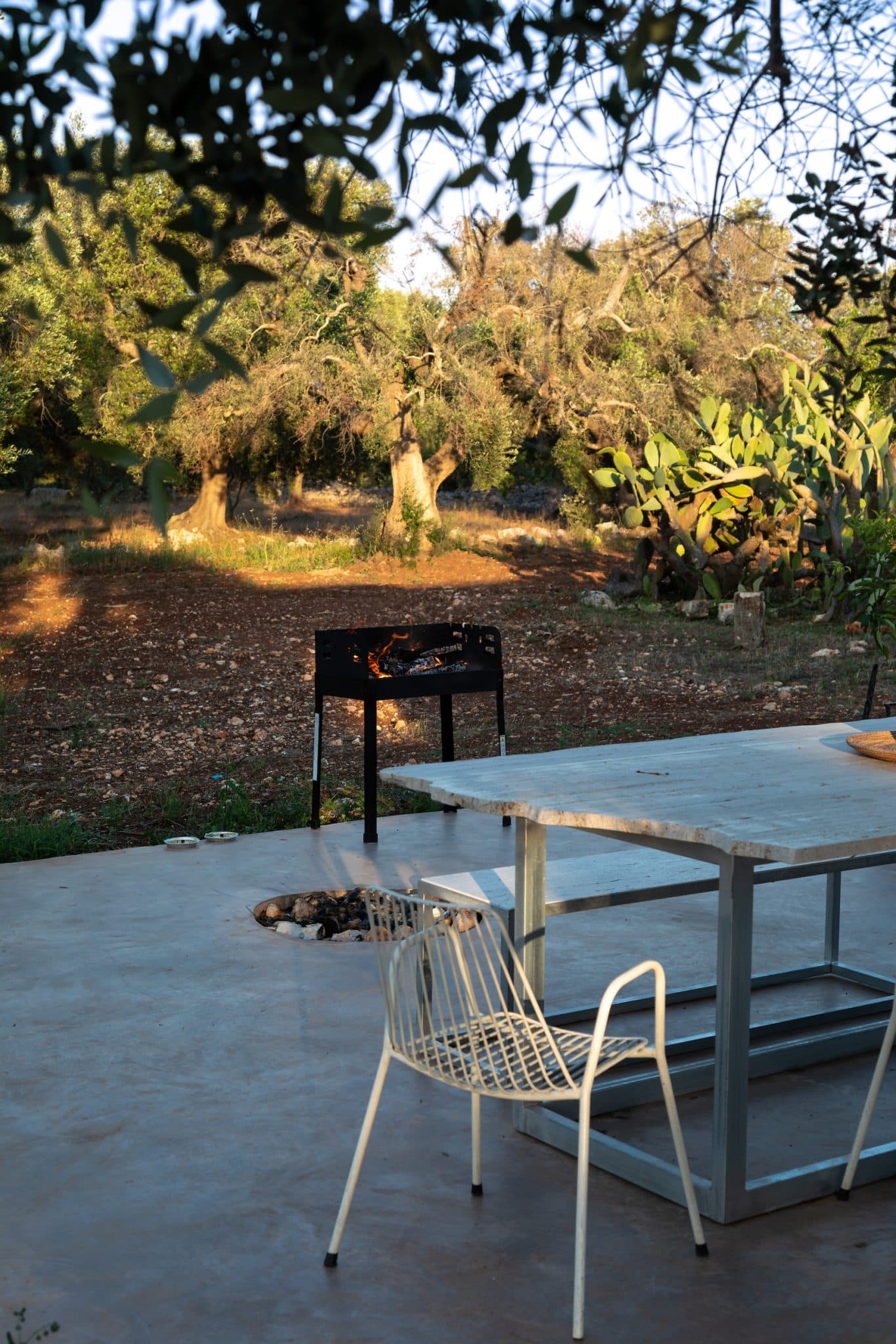 A serene outdoor scene features a sleek concrete patio with a metal table and a white chair. Nearby, a black barbecue with a small fire is set up. The background includes a sunlit, rustic garden with trees and cacti under a clear sky.