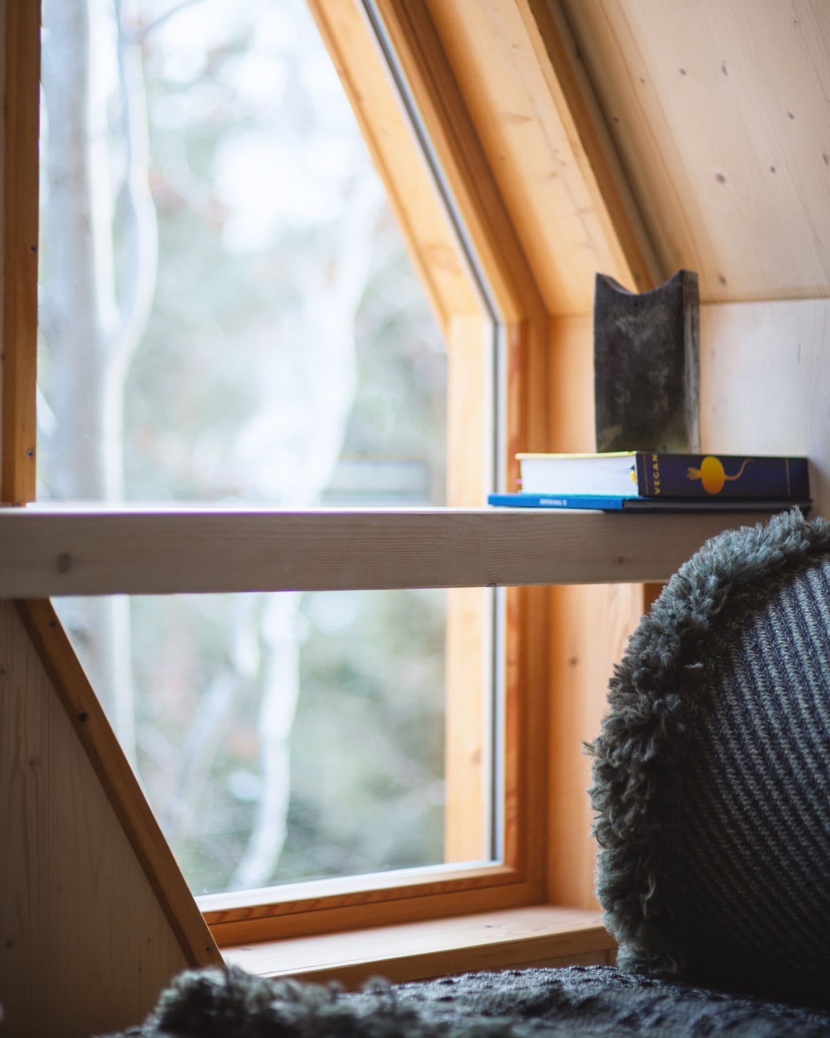 La lumière du soleil filtre à travers une fenêtre triangulaire dans une cabane en bois confortable. Une petite bibliothèque près de la fenêtre contient quelques livres et une sculpture. Un coussin moelleux et texturé est placé sous la fenêtre, créant un coin lecture accueillant. Des arbres sont visibles à l'extérieur.