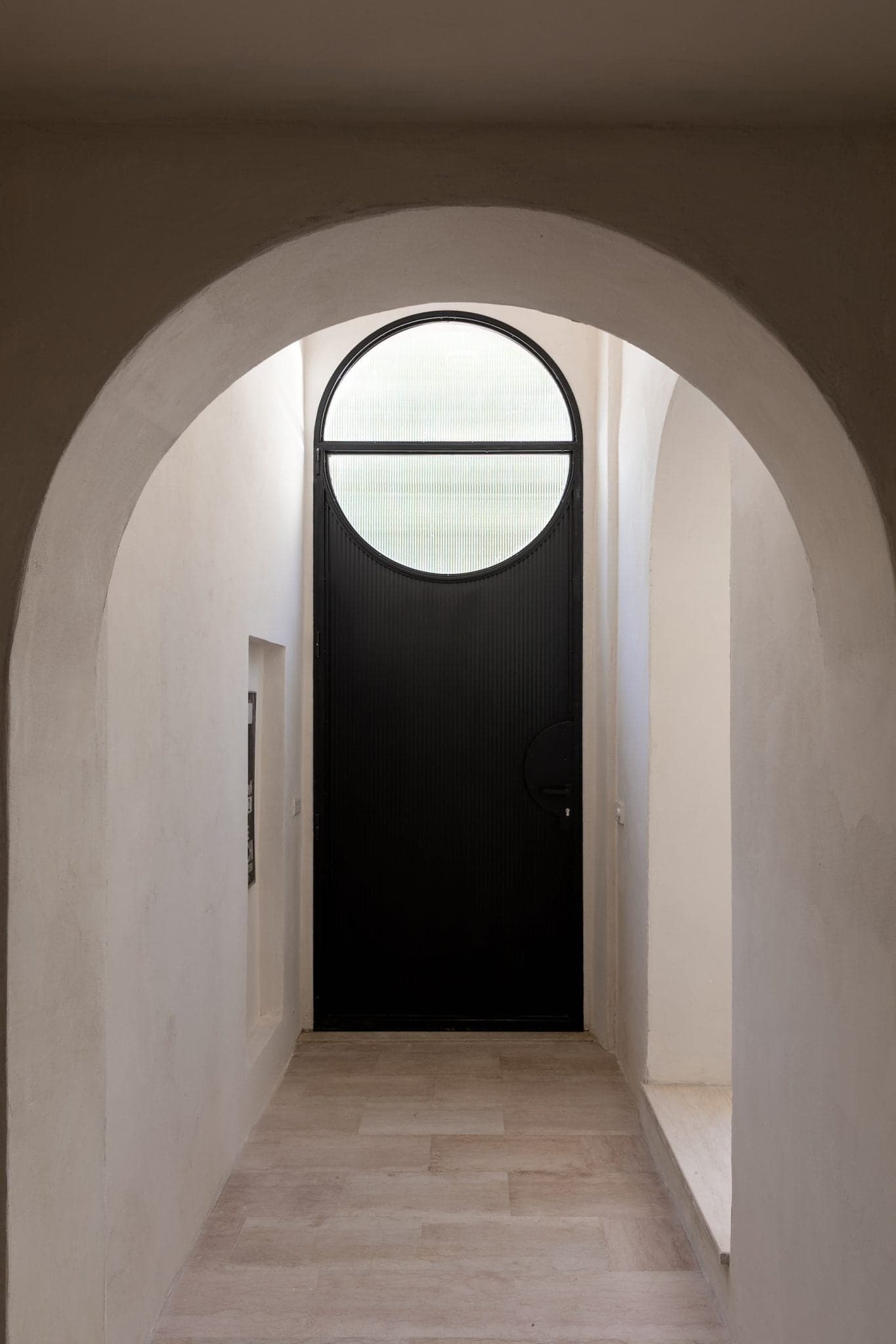 A narrow hallway with light, smooth walls leads to a black door topped with a large circular window. The hallway is illuminated by natural light, and the floor is tiled in beige. An arched doorway frames the view towards the door.