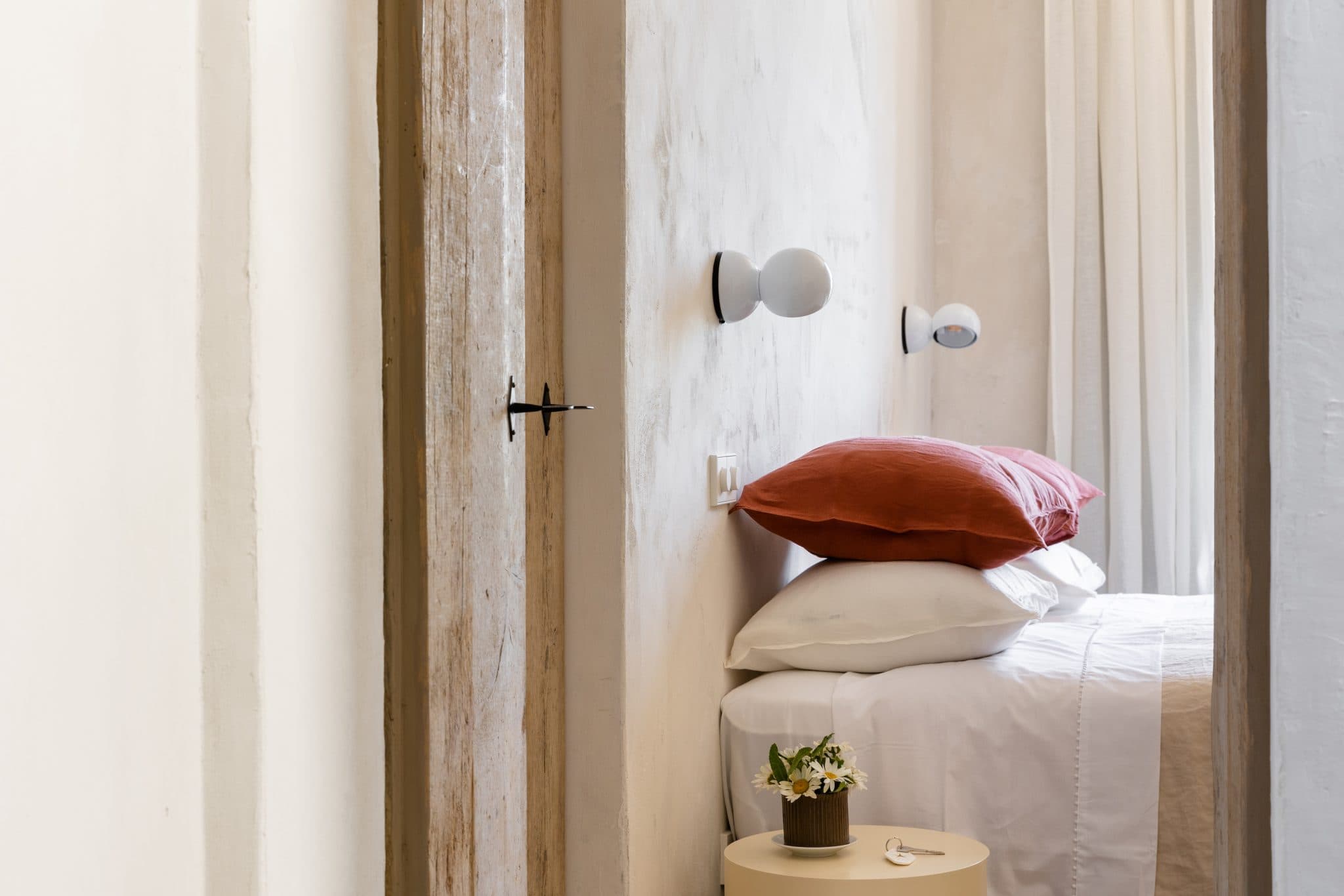 A cozy, minimalist bedroom is visible through a door. The bed is neatly made with white linens and red and white pillows. Two wall sconces are positioned above the bed, and a small round table with a plant decorates the side. The room has a serene atmosphere.