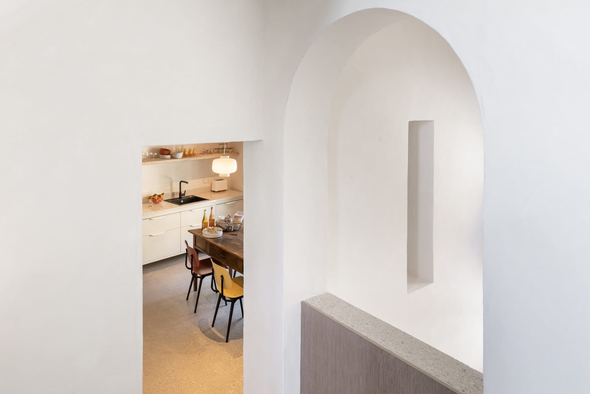 View of a minimalist white kitchen with modern wooden cabinets, a small square window, and a table surrounded by colorful chairs. The foreground features a white arch and a built-in niche, adding architectural interest to the space.