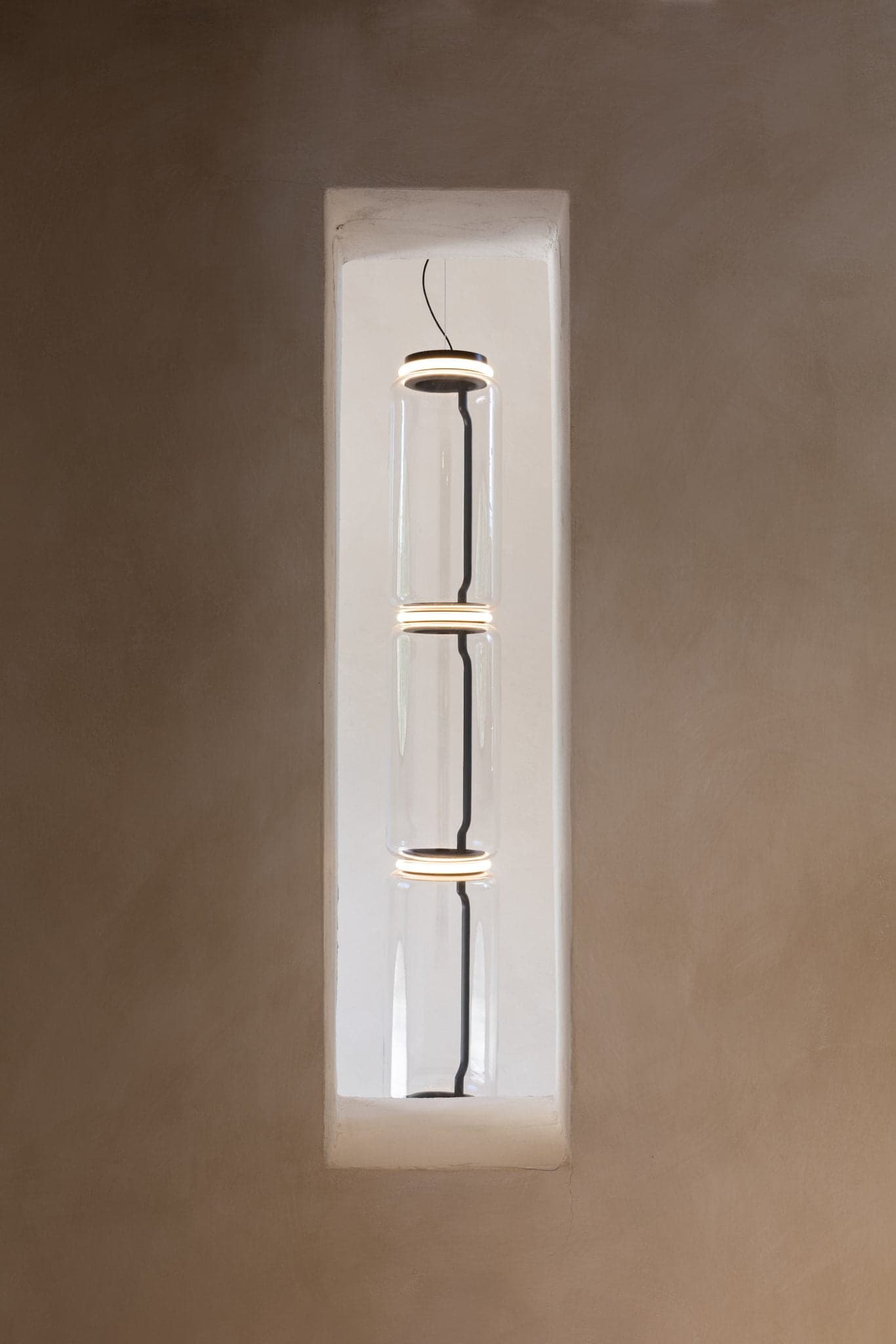 A modern cylindrical glass pendant light, composed of three stacked segments each with illuminated edges, hangs vertically in a narrow, built-in wall niche. The surrounding wall is smooth and painted beige. The design is minimalist and contemporary.