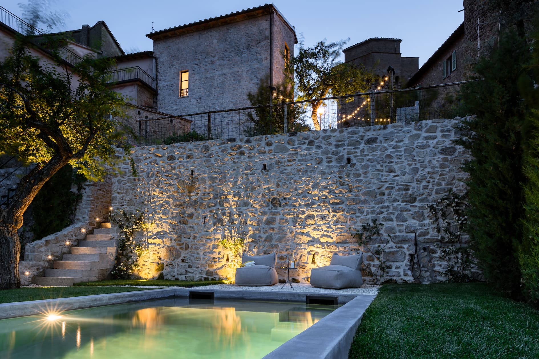 A serene courtyard at dusk with a small illuminated pool surrounded by greenery. Two cushioned seats are placed on a stone patio near the pool, with steps leading up to a stone wall and a lit path. Historical buildings are visible in the background.