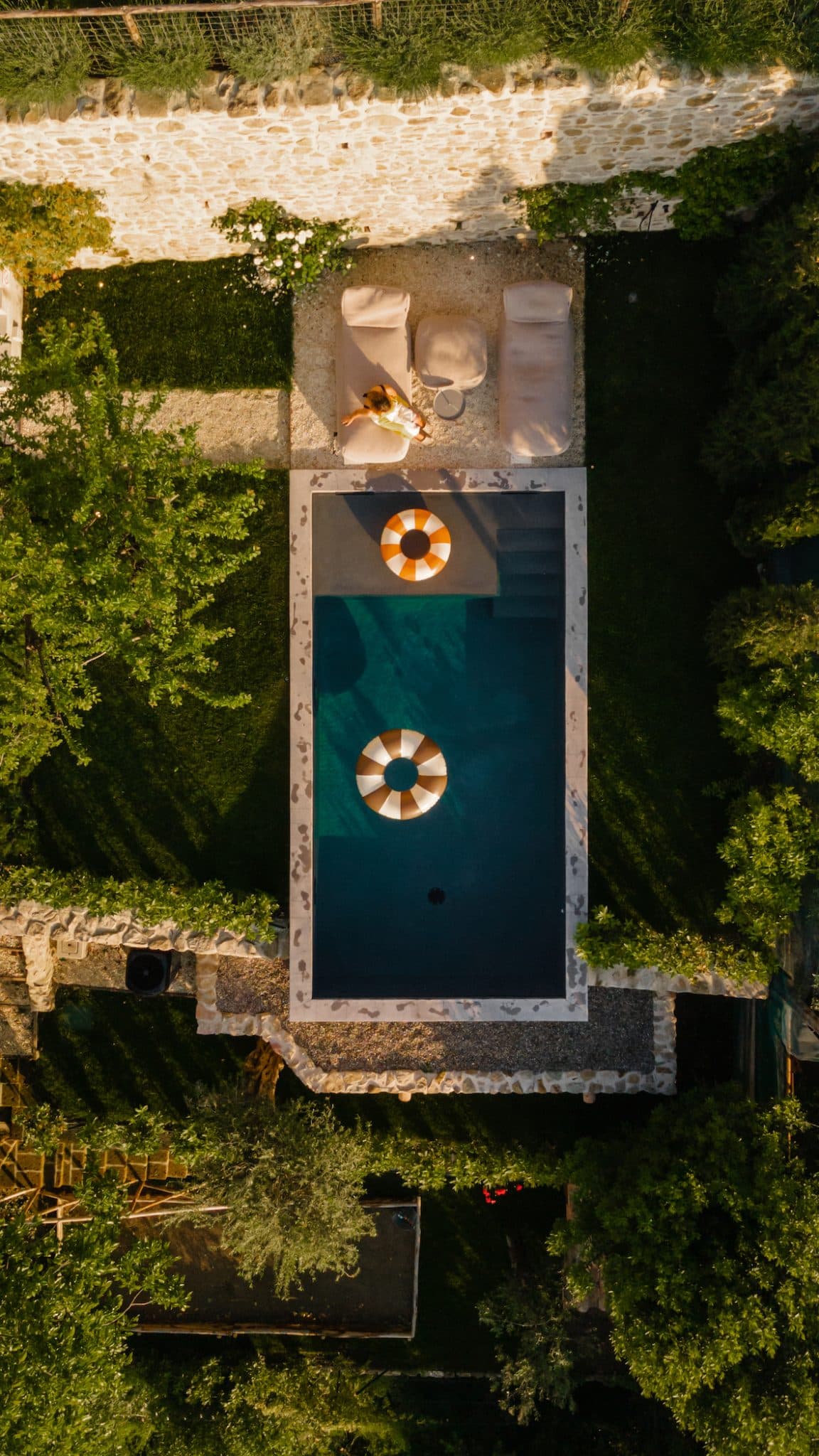Aerial view of a small garden pool surrounded by greenery and a stone wall. Two lounge chairs are positioned at the edge of the pool, with one occupied by a person relaxing. The pool features two colorful inflatable rings floating inside, and a small path leads to the lounge chairs.