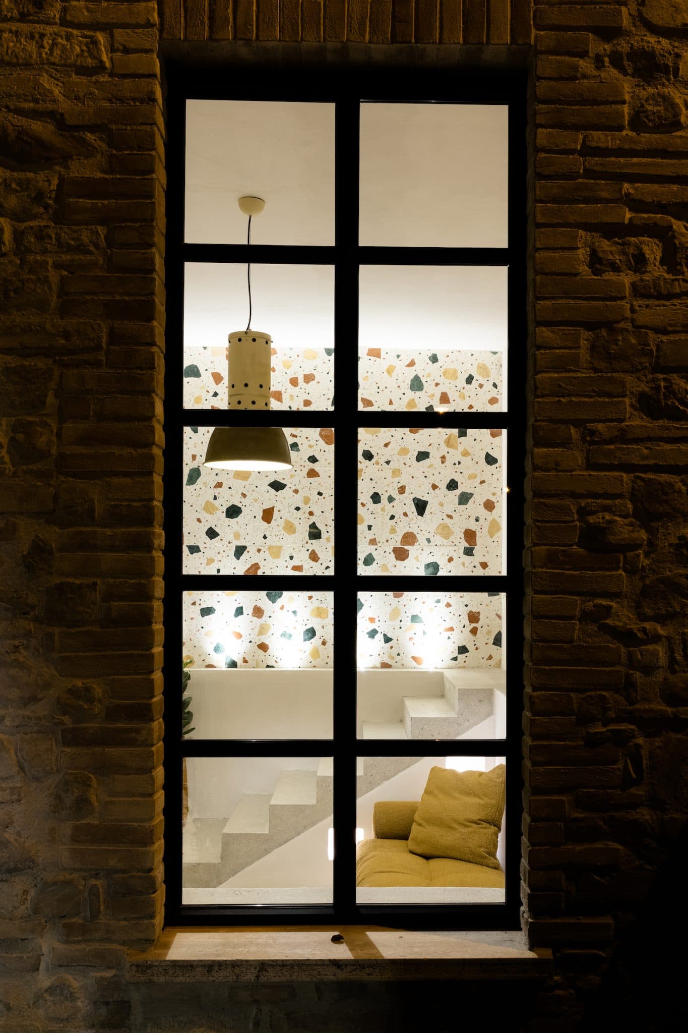 Night view through a large black-framed grid window, revealing a cozy interior. The well-lit room features a terrazzo wall, a hanging lamp, a padded yellow chair, and a staircase with minimalist white steps. The exterior wall is made of raw brown bricks.