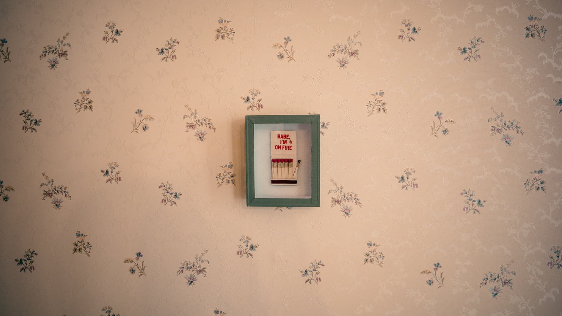 A framed cross-stitch artwork hangs on floral-patterned wallpaper. The frame is green, and the cross-stitch reads "BABE, I'M ON FIRE" with four red and white matches aligned beneath the text. The overall decor has a vintage touch.
