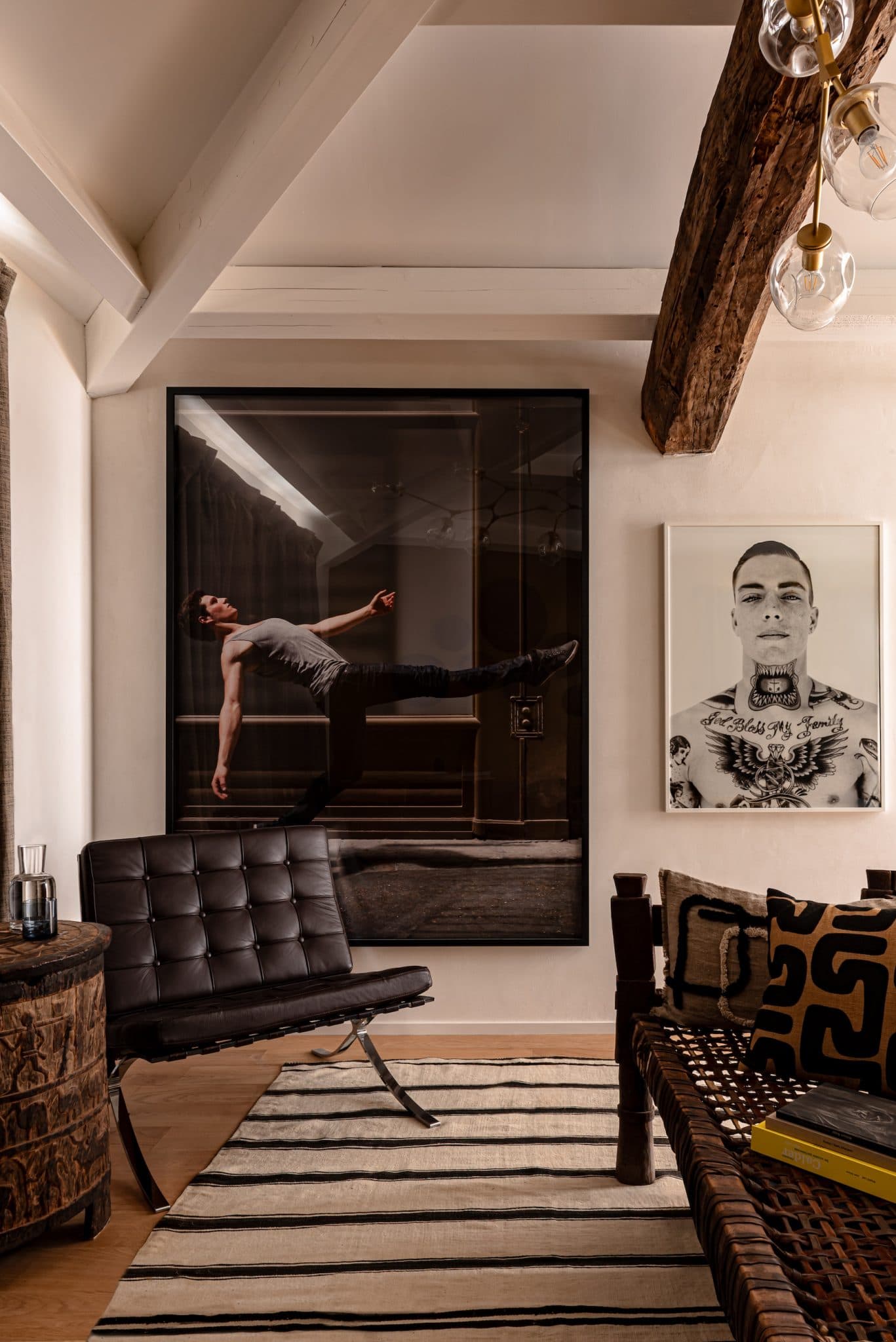 Un salon élégant et moderne avec une grande photographie d'une silhouette flottante sur un mur et un portrait monochrome sur un autre. L'espace comprend une chaise en cuir noir, une table en bois et des coussins à motifs sur un banc. Des poutres en bois apparentes et un tapis rayé ajoutent de la texture.
