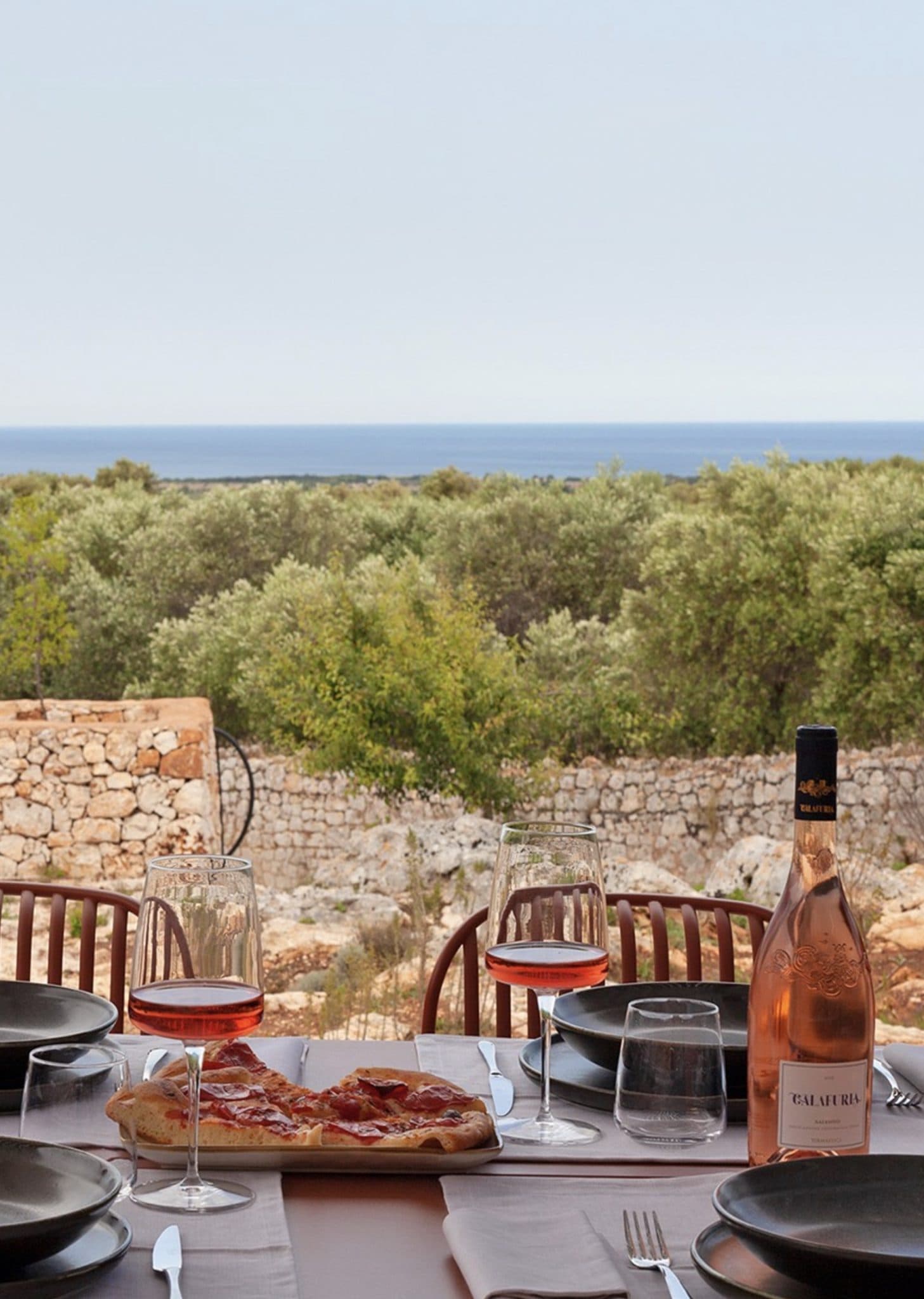 Une table dressée pour un repas avec pizza et verres de vin rosé surplombe un paysage pittoresque d'oliviers et de murs en pierre, avec une vue lointaine sur la mer sous un ciel clair. Des assiettes, des ustensiles et des serviettes sont disposés sur la table, prêts à être servis.