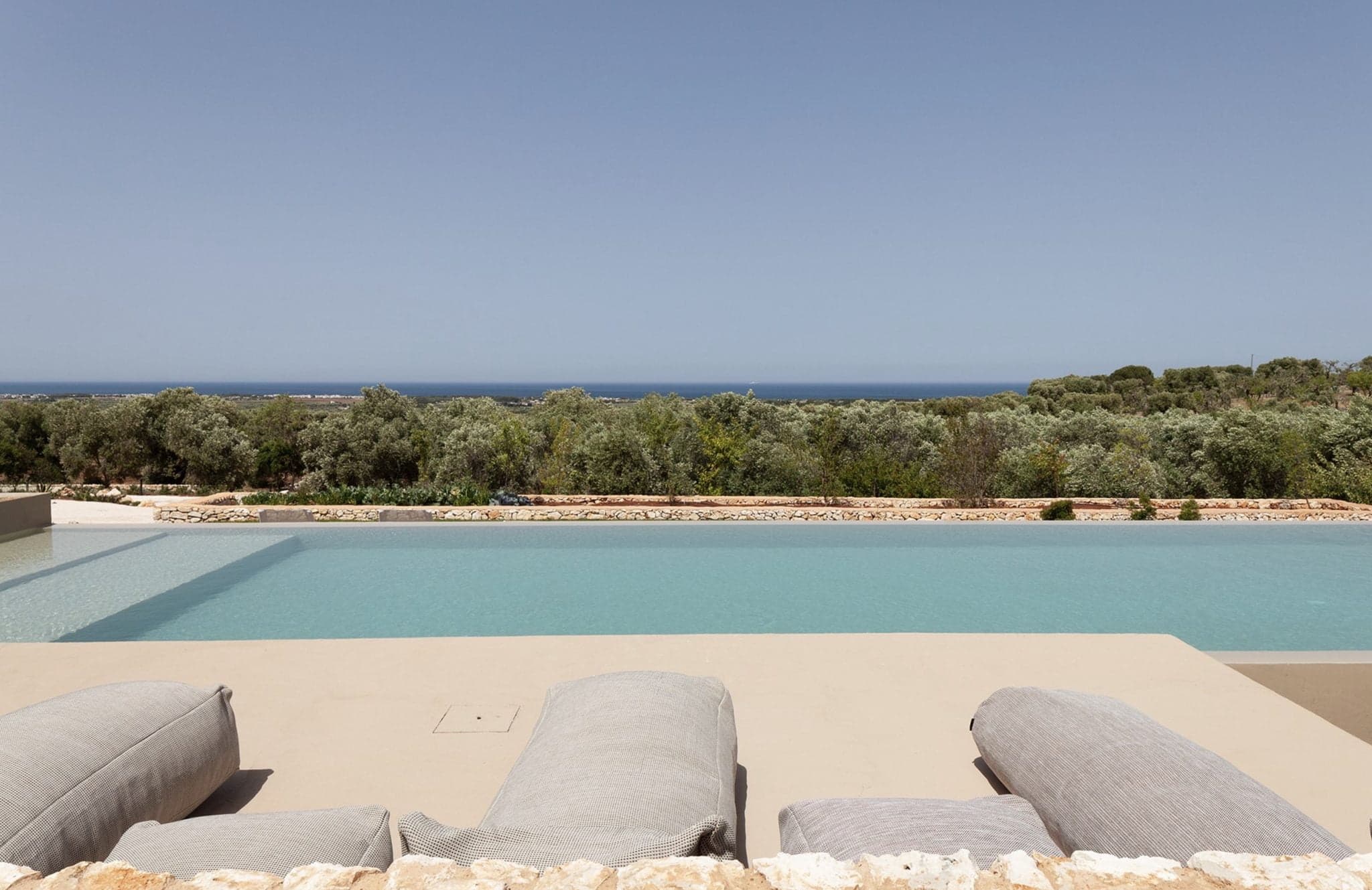 Un cadre extérieur serein comprend une piscine à débordement avec une eau bleu clair, entourée d'un rebord en pierre naturelle. Trois chaises longues grises sont placées au bord de la piscine, offrant une vue tranquille sur la verdure luxuriante et la mer lointaine sous un ciel bleu clair.