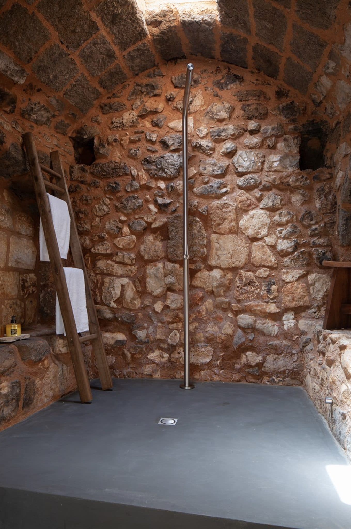 Un espace douche rustique avec des murs en pierre et un seul tuyau de douche en acier inoxydable apparent. À gauche, des étagères en bois de style échelle accueillent des serviettes blanches roulées. Le sol est en ciment lisse et foncé. Le décor dégage une ambiance terreuse et naturelle.