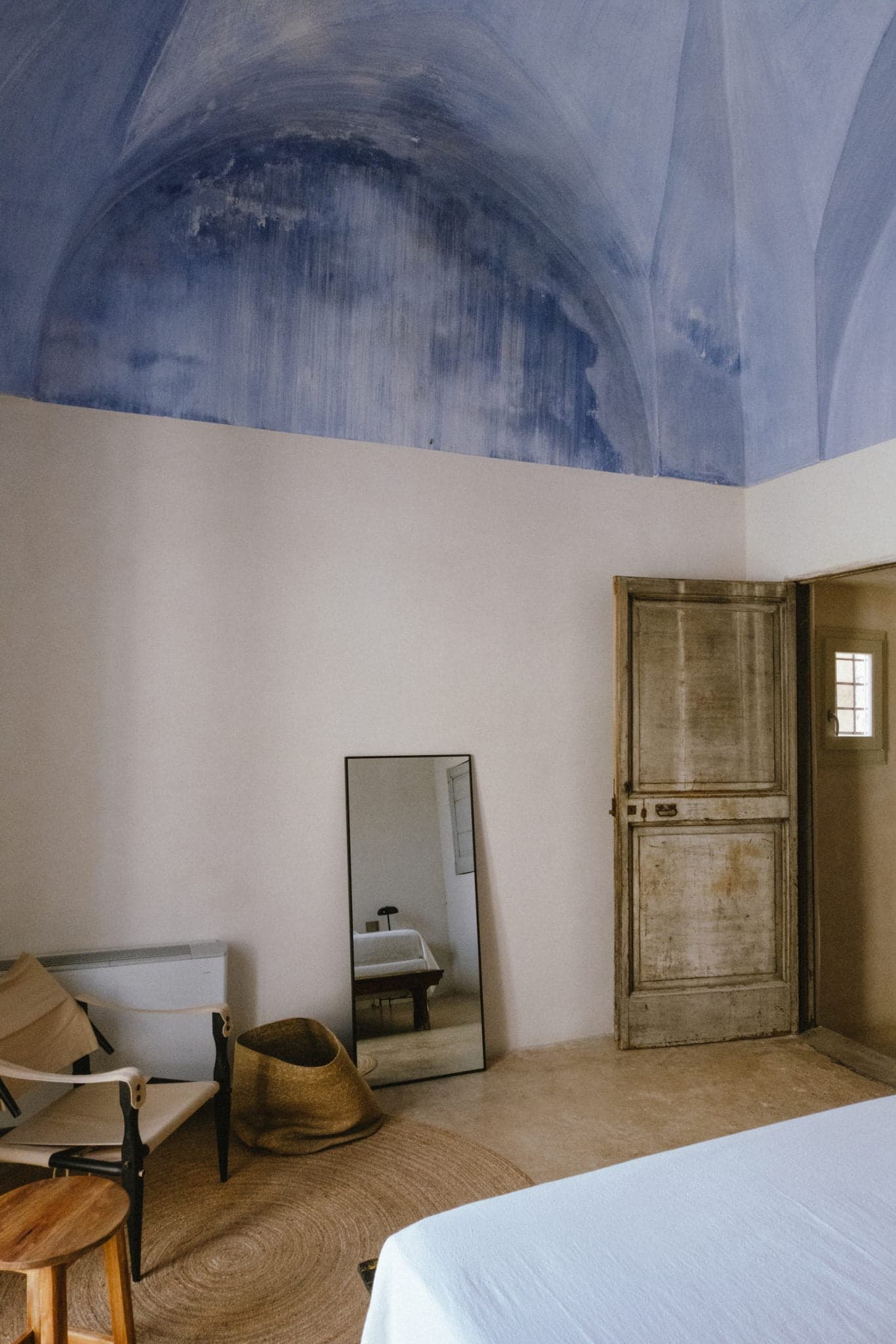 Une chambre minimaliste avec un plafond incurvé peint en bleu. Une porte en bois patiné est légèrement ouverte à côté d'une petite fenêtre. La pièce comprend un miroir appuyé contre le mur, une chaise en bois, un chapeau posé sur un tapis tissé et un lit partiellement visible.