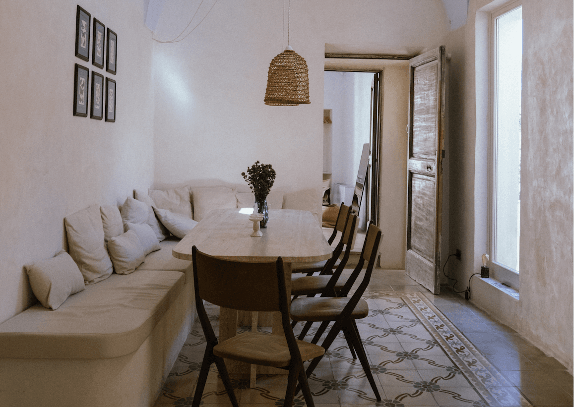 Un coin repas confortable avec un banc blanc rembourré et des chaises en bois autour d'une table en bois clair. Au-dessus de la table est suspendue une suspension en rotin tressé. La pièce est décorée dans des tons neutres, avec des tableaux encadrés sur un mur et des carreaux de sol à motifs. Une porte ouverte révèle un espace lumineux au-delà.