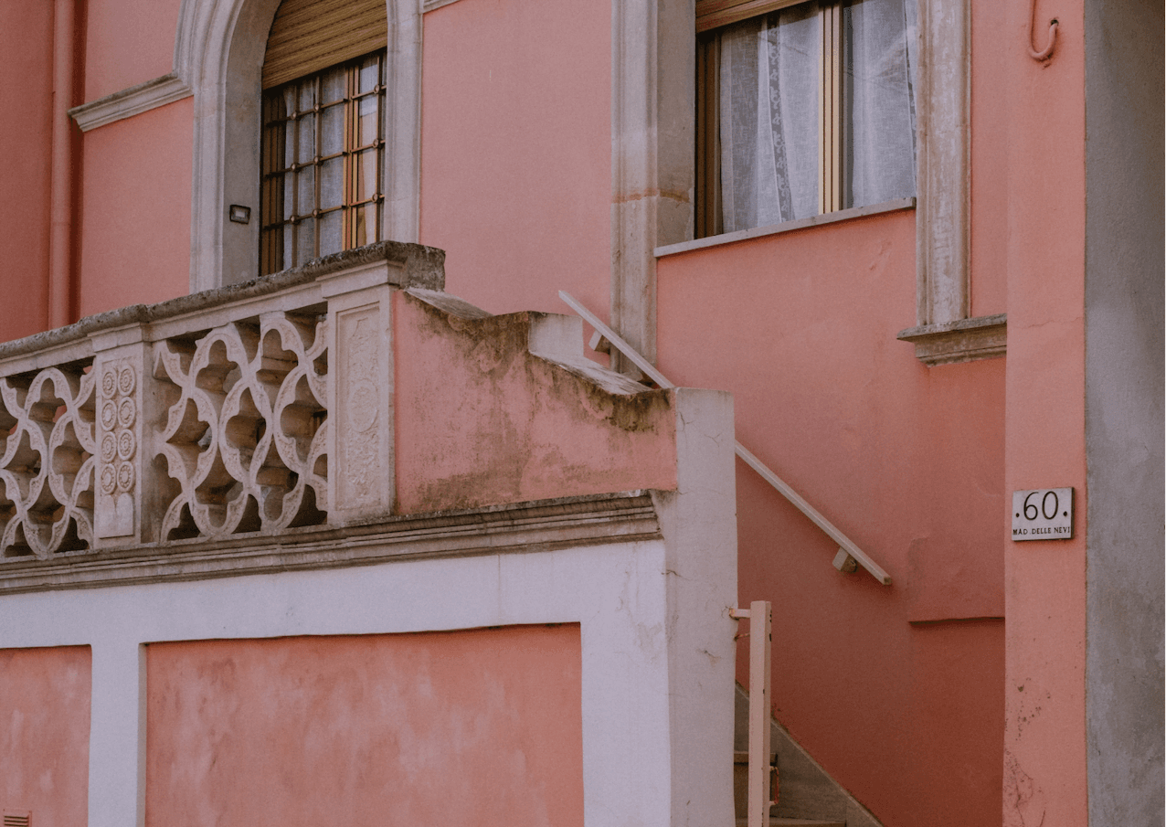 L'extérieur d'un bâtiment peint en rose délavé présente des pierres complexes et patinées sur la balustrade d'un balcon. Un escalier blanc avec une rampe mène à une fenêtre avec des rideaux. Un panneau de numéro de rue « 60 » est fixé au mur à côté de l'escalier.