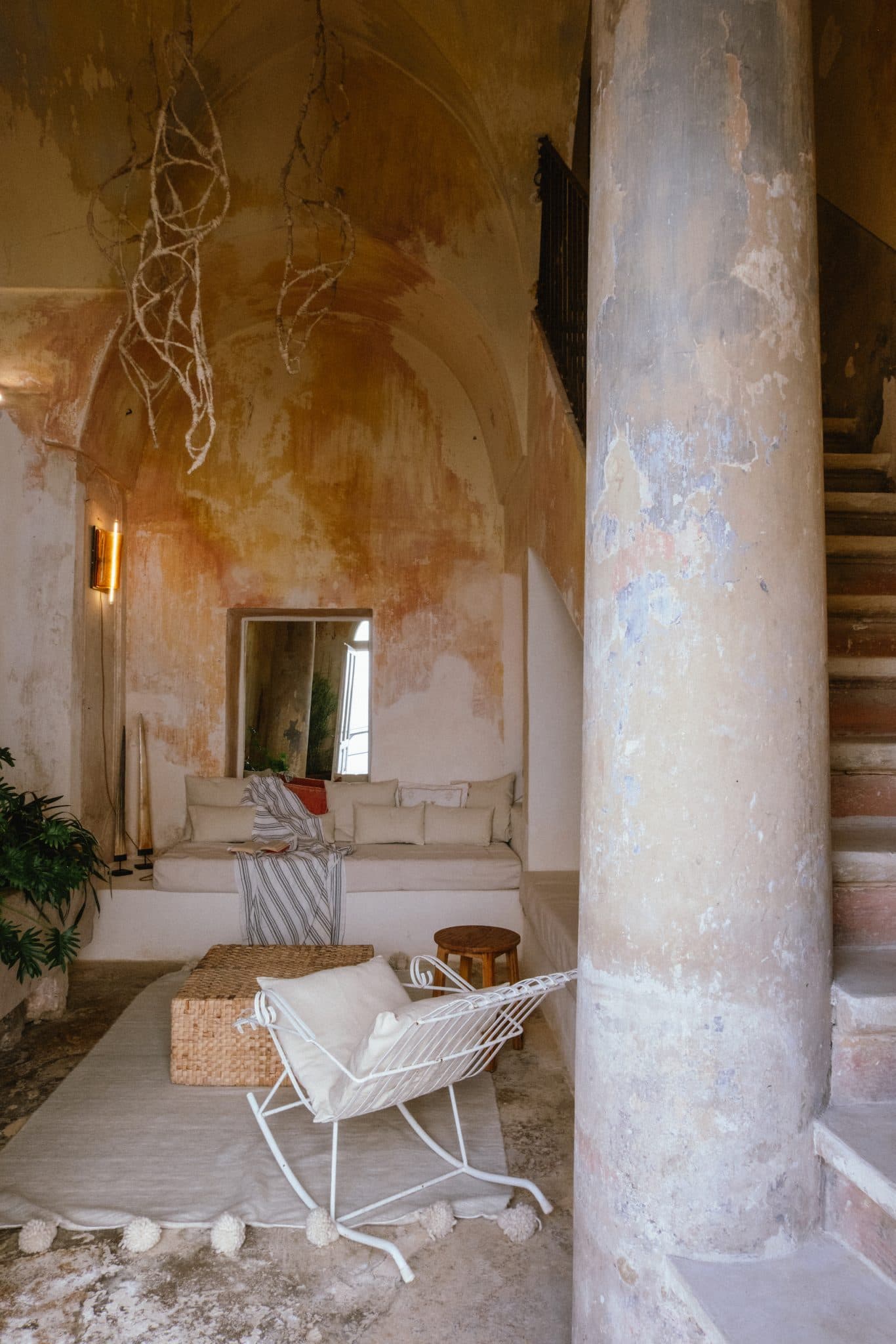 Un espace de vie rustique avec des murs texturés et un coin salon intégré avec coussins blancs, orné de coussins et de plaids. Une chaise en métal blanc fait face à une table en osier et un escalier étroit monte sur le côté droit. Une grande colonne patinée par les intempéries se dresse bien en évidence au premier plan.