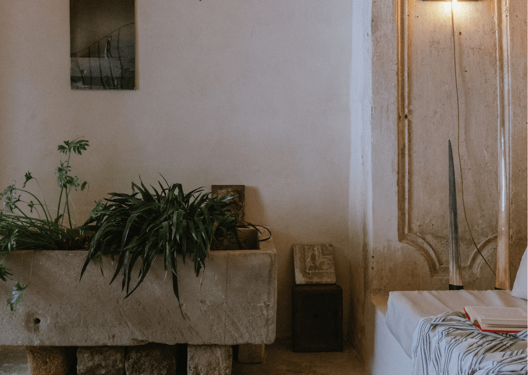 Un coin douillet d'une pièce rustique avec une plante en pot dans un récipient en pierre, un petit banc sous un cadre photo vintage et une épée accrochée au mur. Un lit ou un canapé blanc avec un livre et une couverture rayée est partiellement visible à droite. Un éclairage doux crée une atmosphère chaleureuse.
