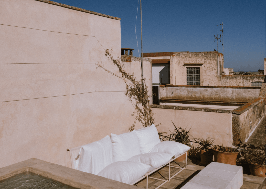 Une terrasse sur le toit avec un petit canapé d'extérieur orné de coussins blancs. Les murs beiges des bâtiments environnants sont visibles sous un ciel bleu clair. Une plante grimpante verte grimpe sur un mur et des plantes en pot sont placées près du canapé.