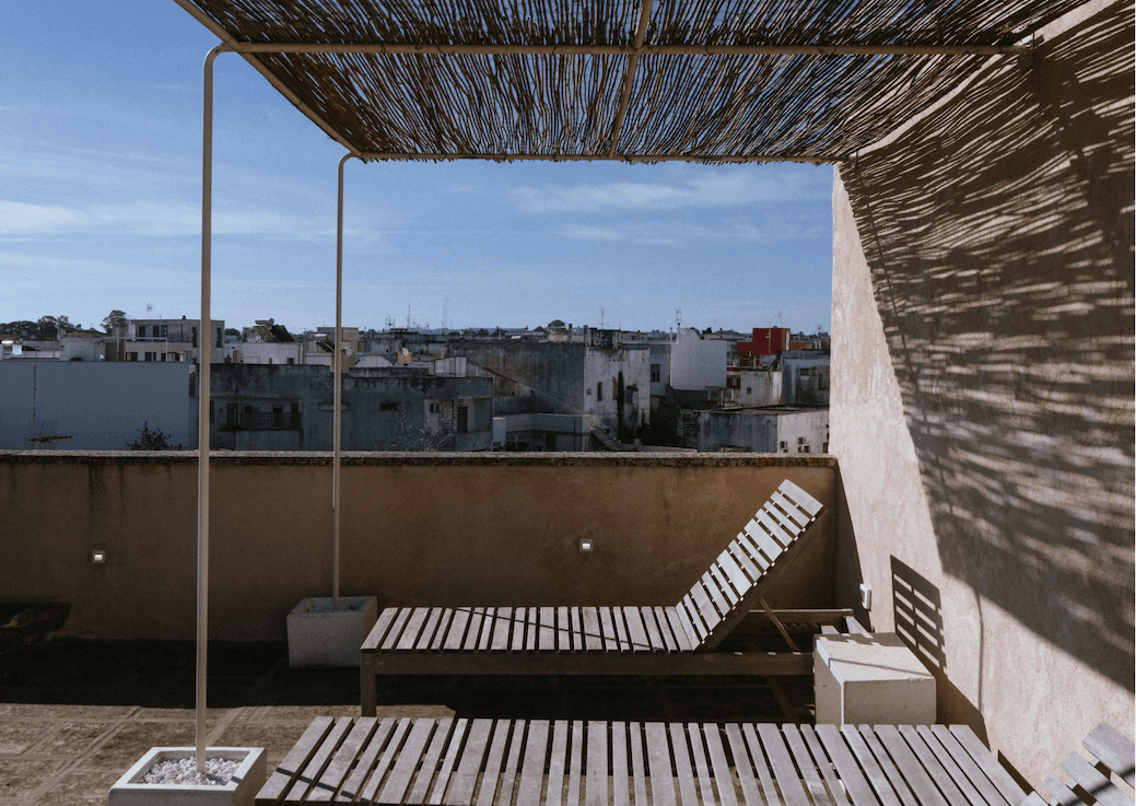 Une terrasse sur le toit avec des chaises longues en bois et une pergola ombragée projetant des ombres rayées. La vue donne sur une ville aux bâtiments blancs sous un ciel bleu clair.