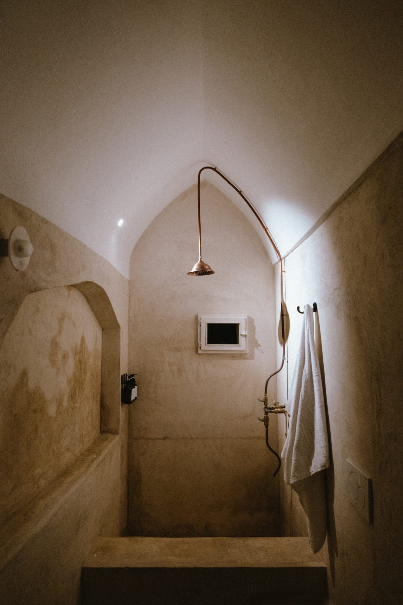 Une salle de bains minimaliste au style rustique comprend un espace douche en pierre et une pomme de douche incurvée en cuivre qui s'étend du plafond. Une petite fenêtre et une serviette sur un crochet sont visibles sur les murs texturés beiges, éclairés par un éclairage d'ambiance doux.