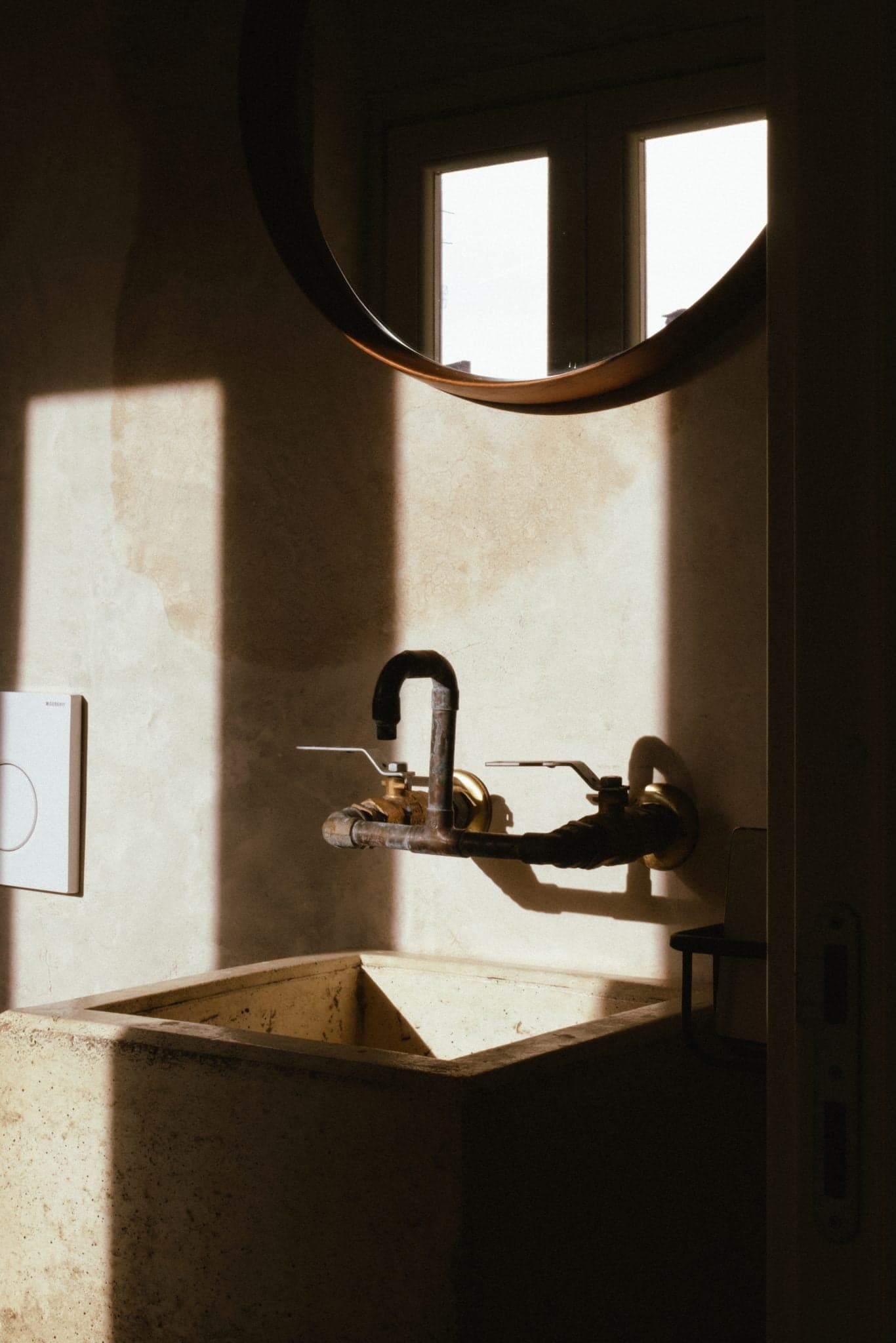 Une salle de bain faiblement éclairée avec un miroir rond au-dessus d'un lavabo en pierre beige. Un robinet à l'ancienne est fixé au mur au-dessus du lavabo. La lumière du soleil pénètre par une fenêtre, projetant des ombres sur les murs texturés. Un objet fixé au mur est partiellement visible à gauche.