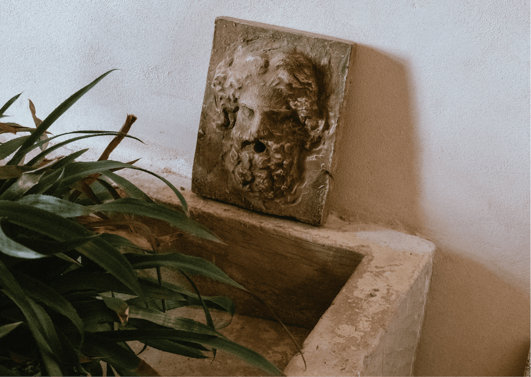 Un relief en pierre vieillie représentant un visage barbu est fixé au mur au-dessus d'un bassin en pierre, avec des plantes à feuilles vertes recouvrant partiellement le côté gauche. L'arrière-plan présente un mur lisse et blanc cassé. L'ambiance générale suggère un cadre antique ou rustique.