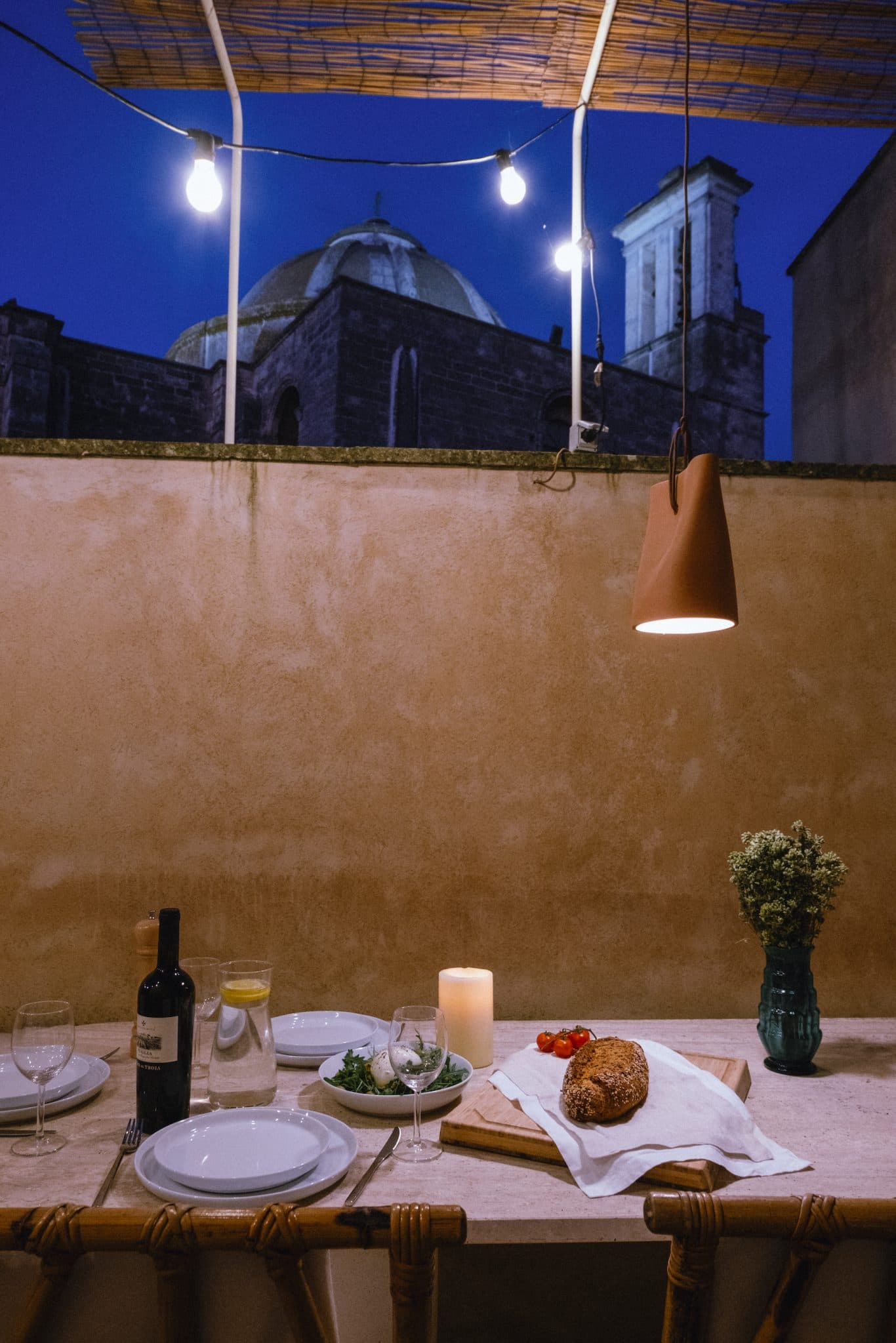 Une table de dîner en plein air confortable avec une miche de pain, une salade et une bouteille de vin. La table est ornée d'une bougie et d'un bouquet de fleurs. Des guirlandes lumineuses suspendues illuminent le décor et, à l'arrière-plan, un dôme et une tour éclairés sont visibles dans le ciel du soir.